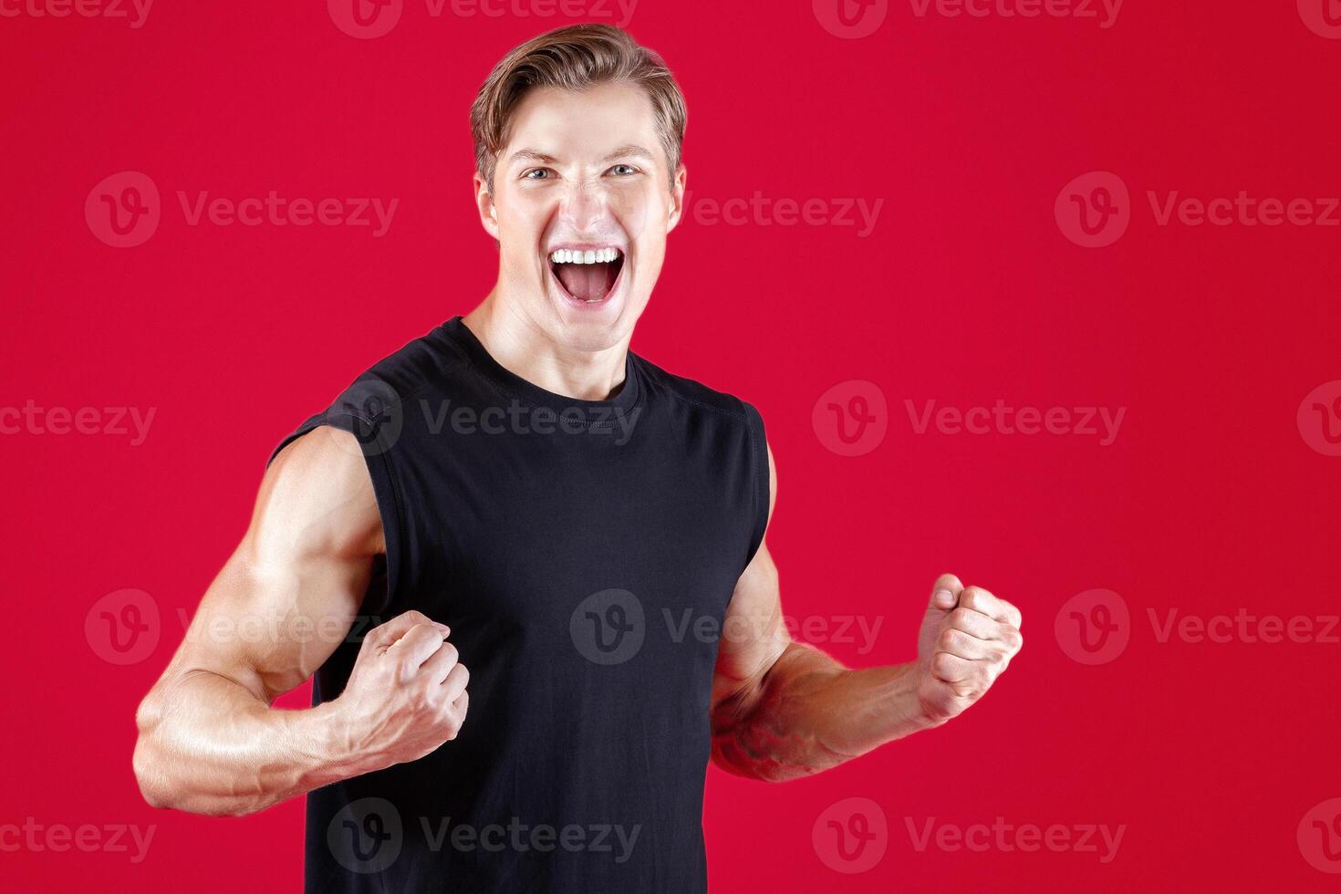 Emotional Caucasian sportsman screaming, making YES gesture, celebrating his victory on red studio background. Triumphant young bodybuilder winning competition, taking first place in sports contest photo