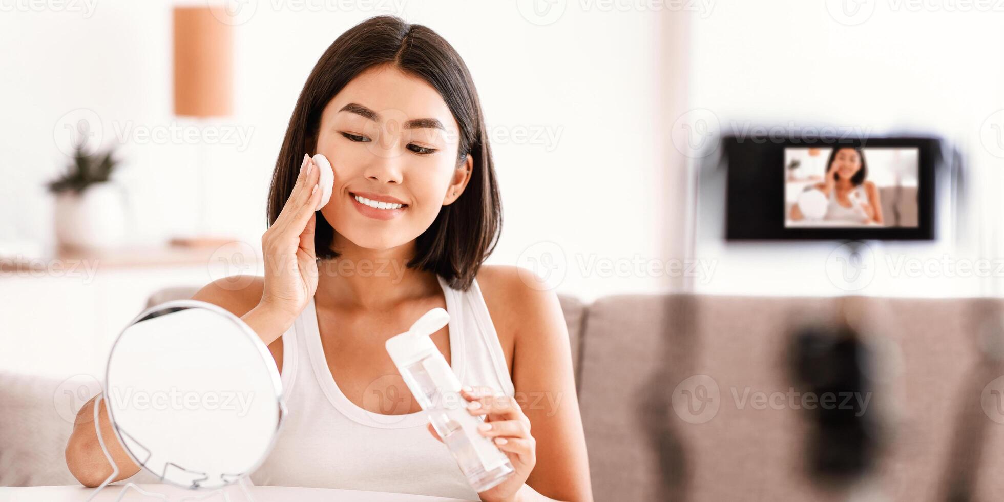 Applying Toner. Asian Woman Cleaning Face With White Pad Looking In Mirror. Beautiful Lady Removing Makeup With White Cosmetic Cotton Pad, Recording Vlog On Camera For Social Media Channel photo
