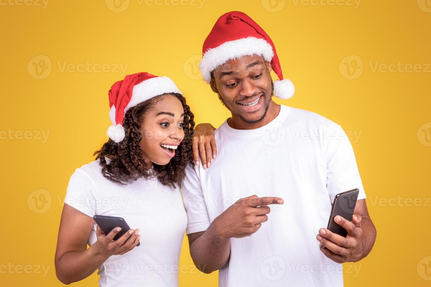 Christmas Mobile App. Excited Black Couple Holding Cellphones Recommending App For Smartphone, Posing Wearing Santa Hats Over Yellow Studio Background. Xmas Advertisement. Panorama photo