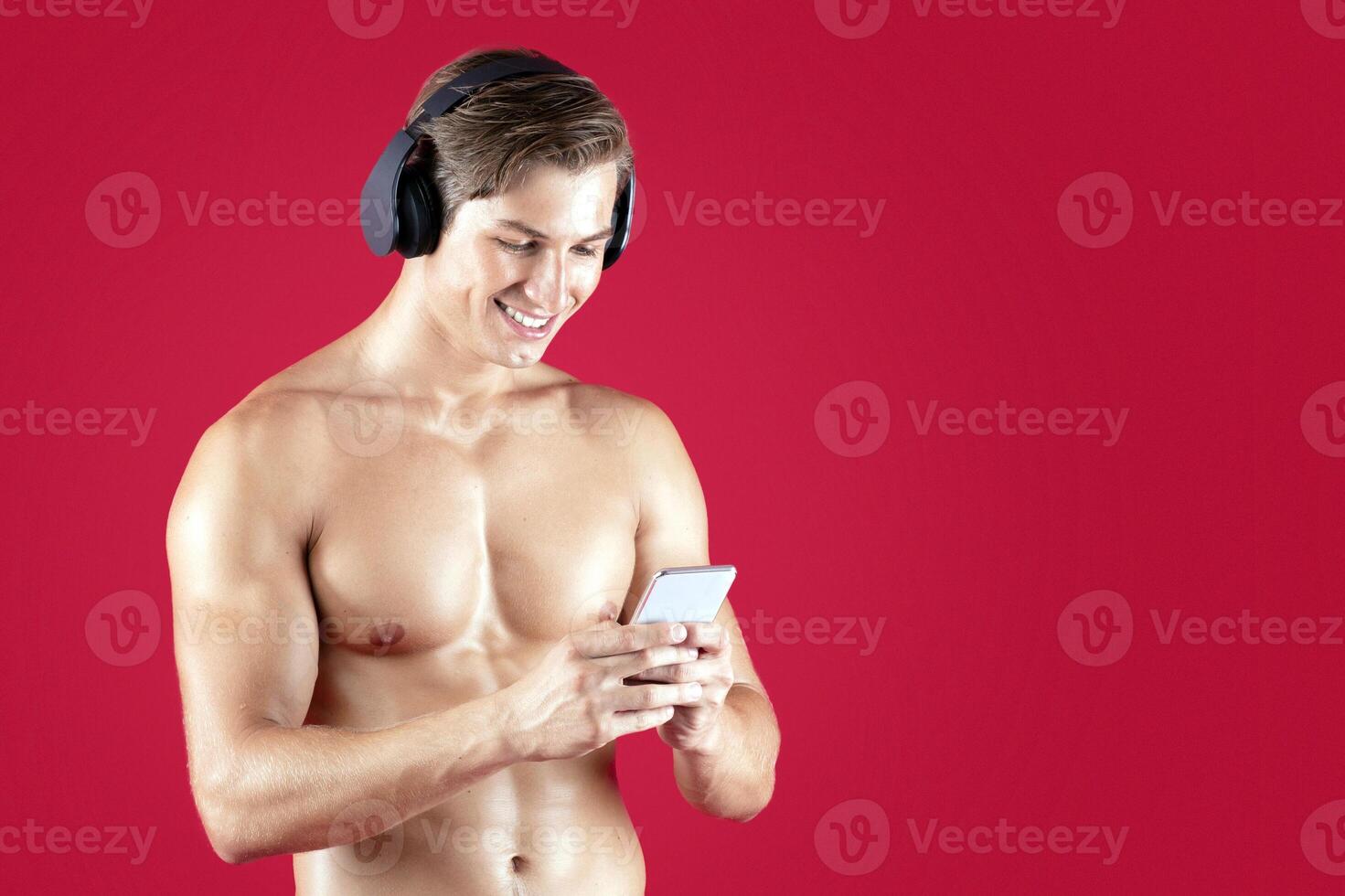 Handsome young athlete with bare torso listening to music in headphones, choosing song on cellphone over red studio background. Muscular millennial bodybuilder picking soundtrack for workout photo