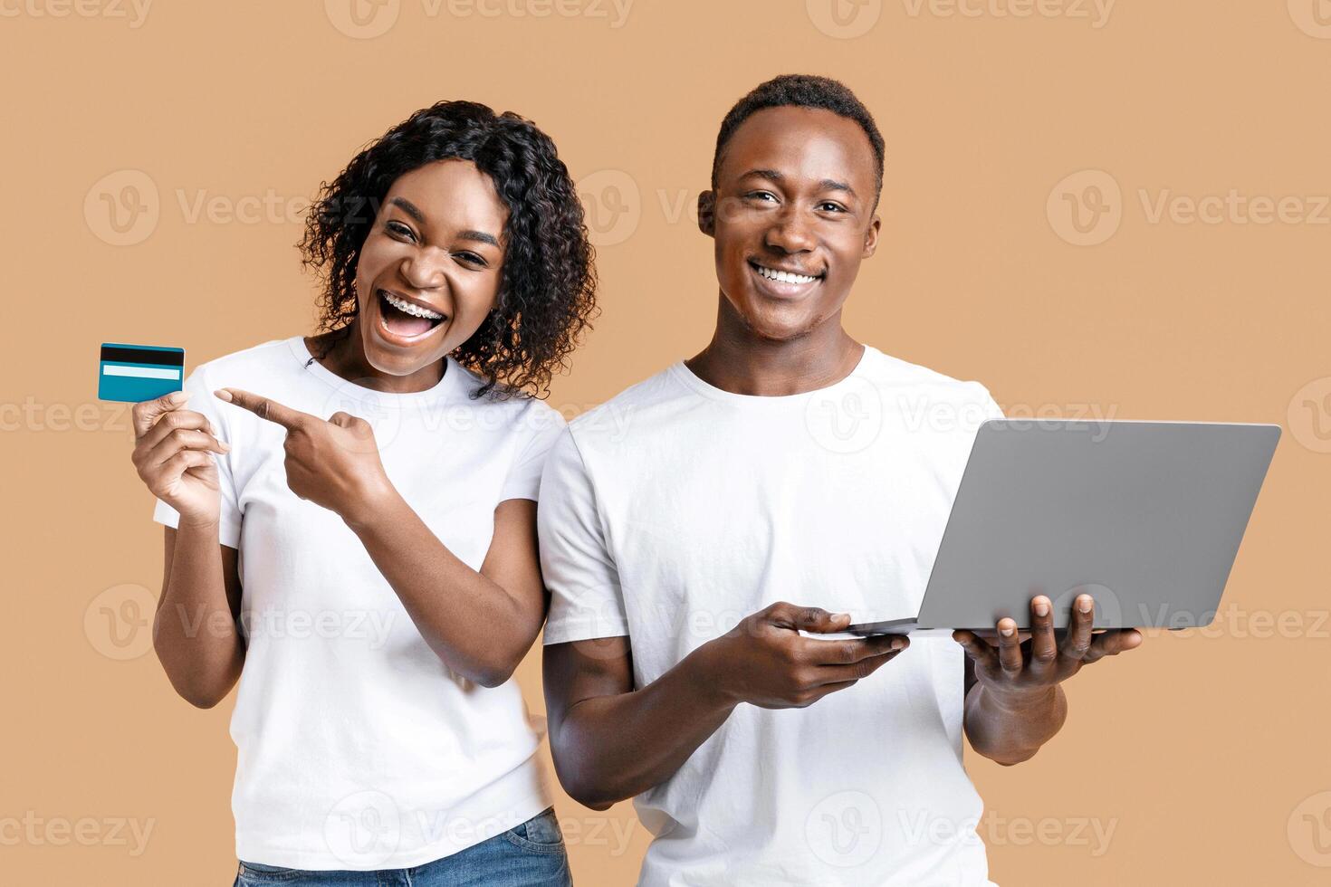 Online banking and e-commerce. Happy african-american man and woman pointing at credit card and working on laptop on yellow studio background. Black couple making purchases online, using laptop photo