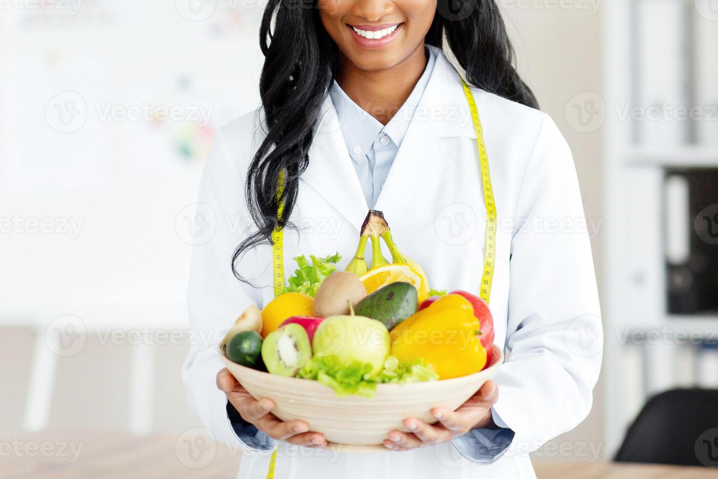 Portrait of joyful black nutritionist in lab coat holding bowl of fresh fruits and veggies at weight loss clinic. Healthy nutrition consultant recommending balanced plant based diet photo