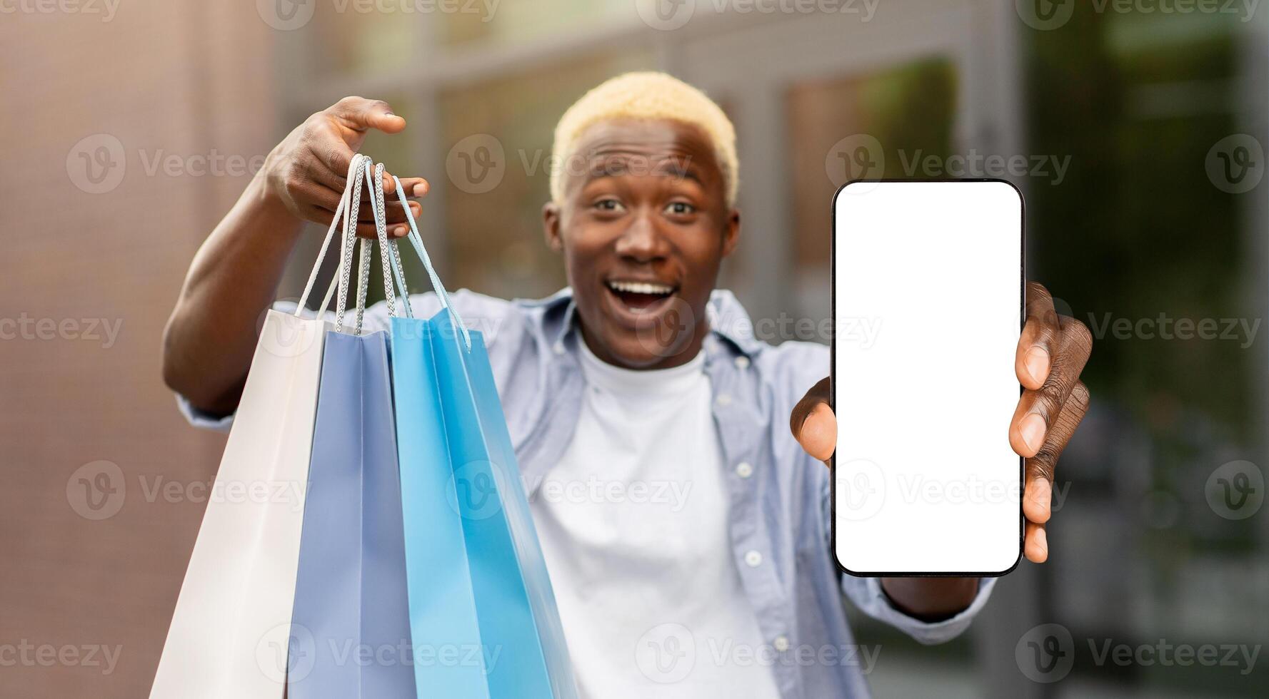 Unbelievable bonus for mobile user, online shopping application. Surprised african american man with packages shows smartphone with blank screen to camera photo