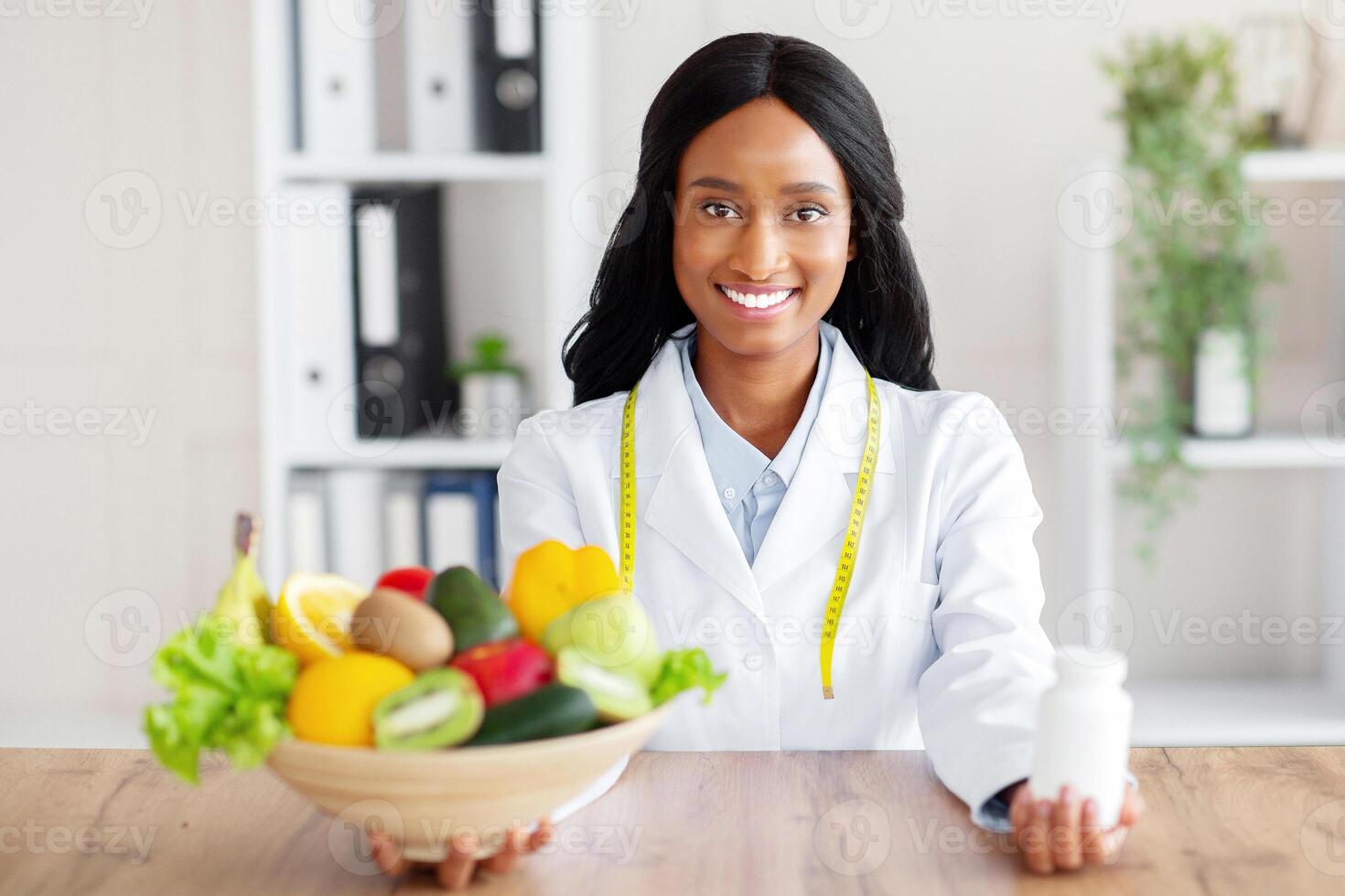 Portrait of black female dietitian holding bowl of fresh products and jar of tablets at table in clinic. Young nutritionist offering choice between healthy eating and medications. Healthcare concept photo