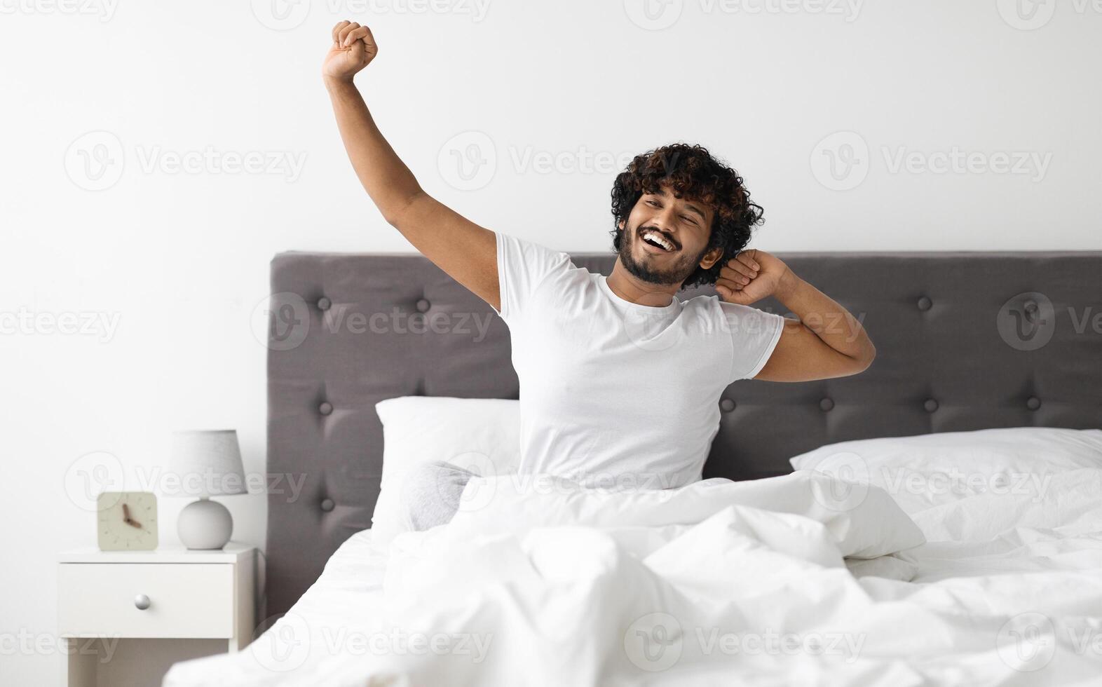 Well-rested happy young indian guy with beard and curly hair sitting on bed and stretching, enjoying new day, looking at copy space and smiling, cozy bedroom interior. Healthy sleep concept photo