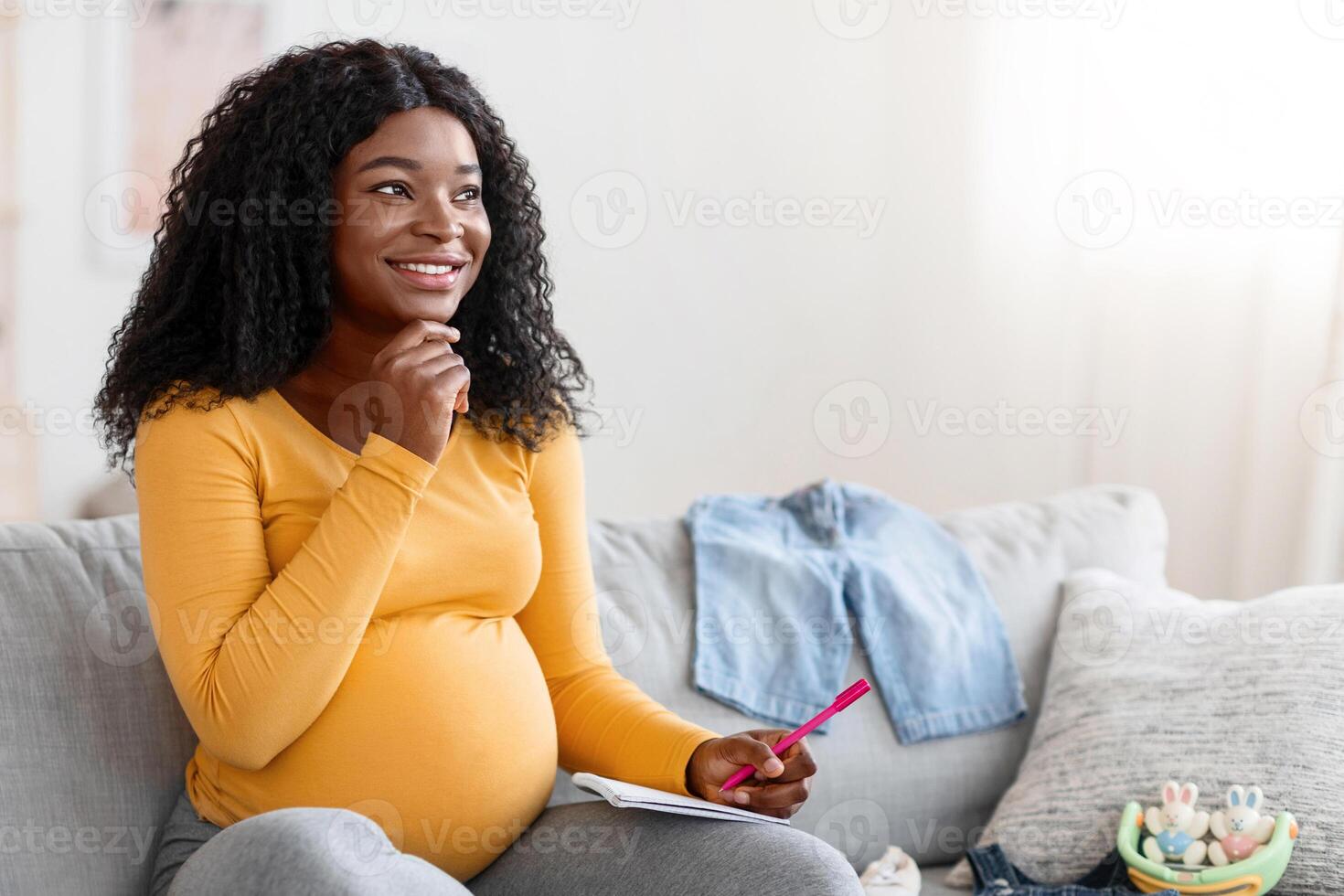 Excited pregnant black woman sitting on sofa with notebook and pen, making shopping list before delivery, free space. Pensive african american mother getting ready for baby coming, home interior photo
