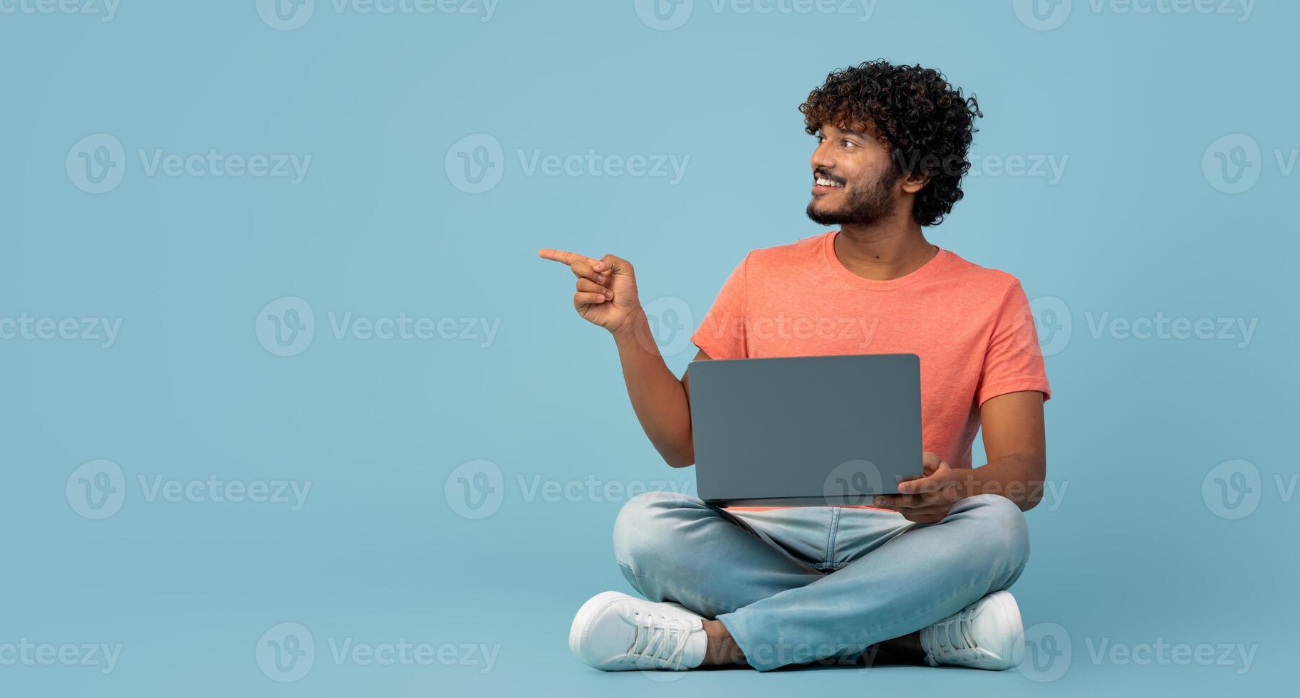 Handsome millennial indian man in casual outfit sitting on floor, using modern laptop on blue studio background, typing on computer keyboard, working online, pointing at copy space, panorama photo