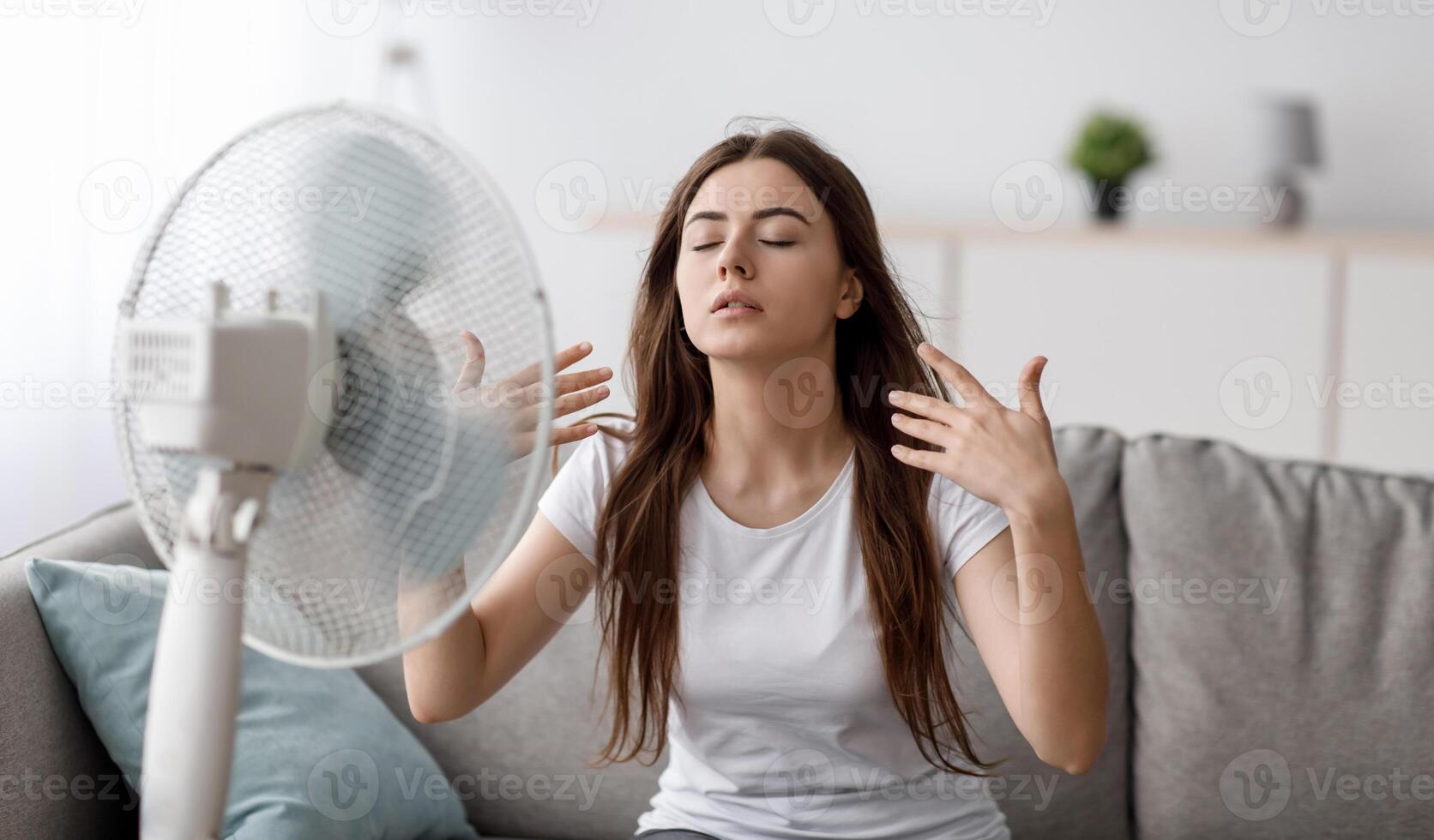 Sad young european woman suffering from heat sits on sofa, catches cold air from fan and waves hands to herself in living room interior. Summer weather and overheating without air conditioning at home photo