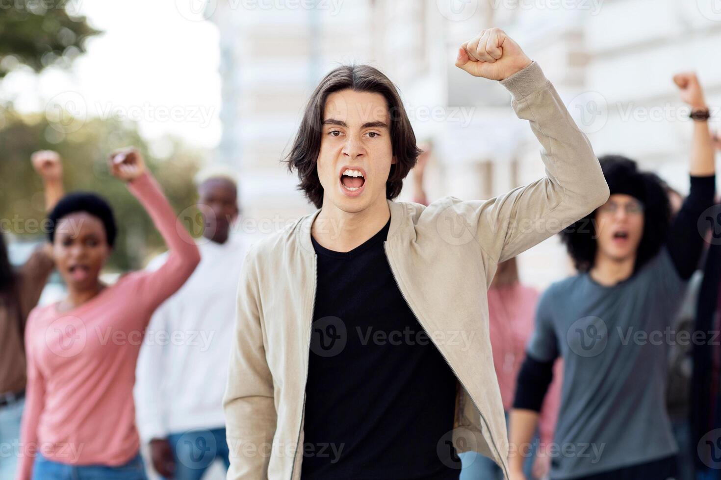 Multiracial group of activists chanting slogans, raising fists up, making strike on the street. Male leader guiding millennial men and women different nationalities, protesting against discrimination photo