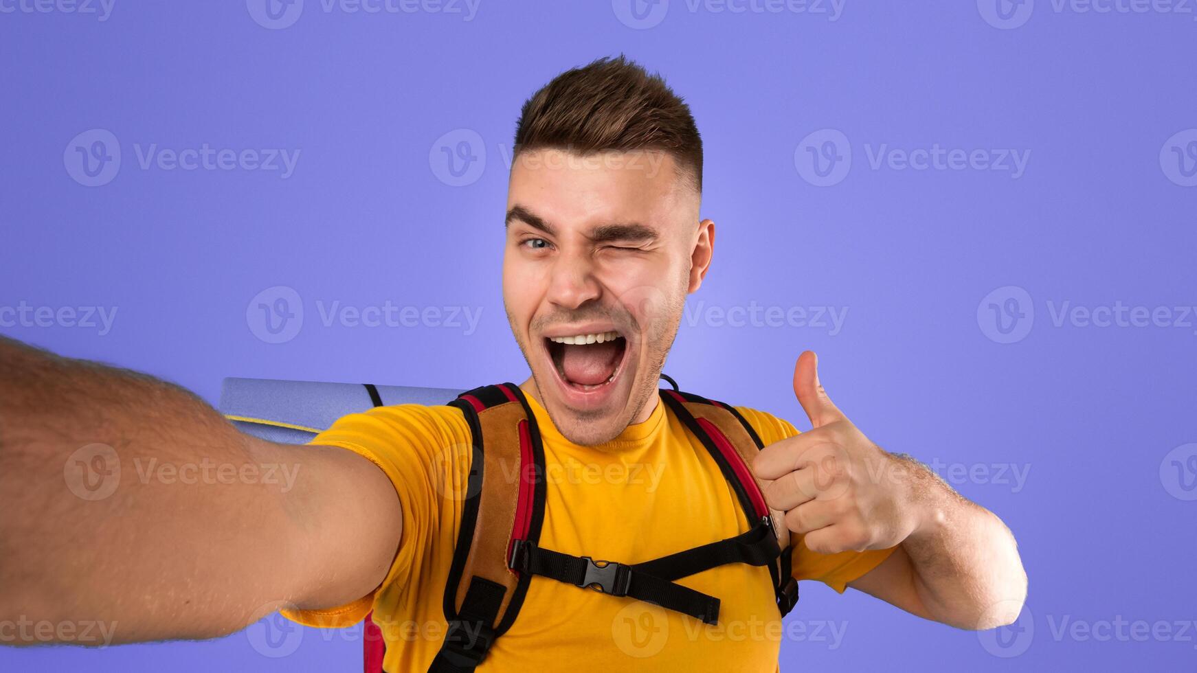 Cool young hipster with tourist equipment taking selfie, winking and showing thumb up gesture over blue studio background. Positive hiker taking funny photo of himself before camping trip