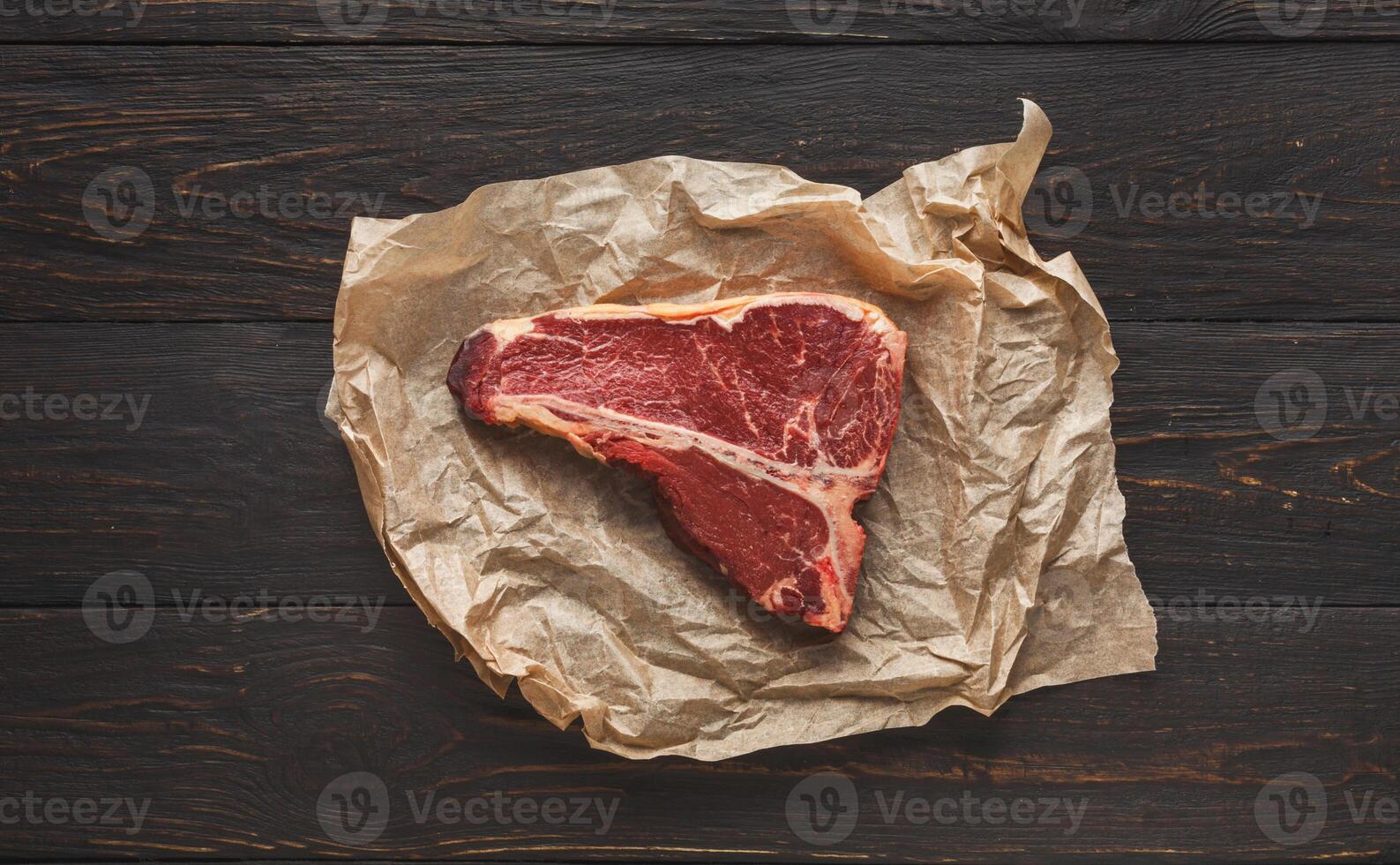 Raw t-bone steak on craft paper on dark background. Prime meat on black table with spices, copy space, top view photo