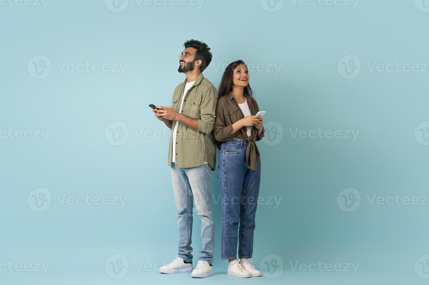 Dating mobile app concept. Stylish young eastern man and woman hipsters standing back to back, using smartphones on colorful studio background, chatting online, looking at copy space, full length photo
