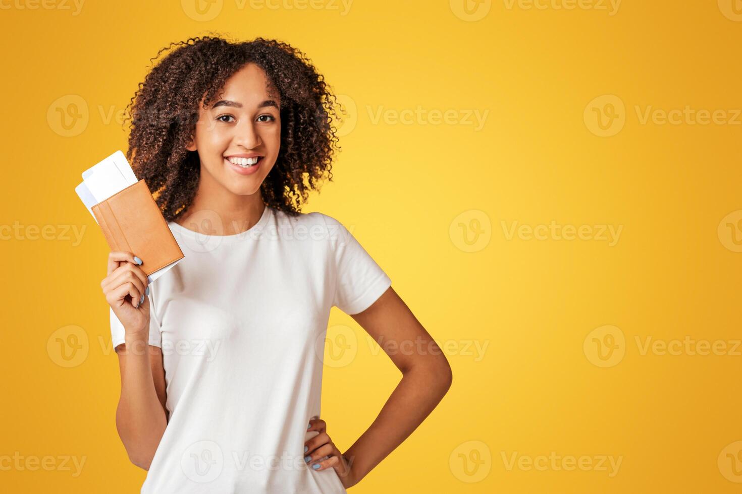 Traveler and vacation. Young with snow-white smile african american lady with curly hair in white t-shirt holding passport with air tickets, isolated on yellow background, panorama, empty space photo