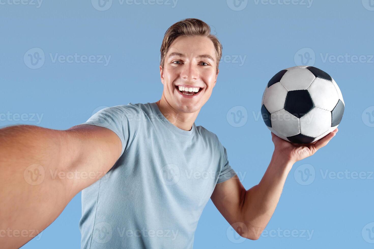 Smiling young man with soccer ball taking selfie on his mobile phone over violet studio background. Positive football fan taking photo of himself before championship, making good memories