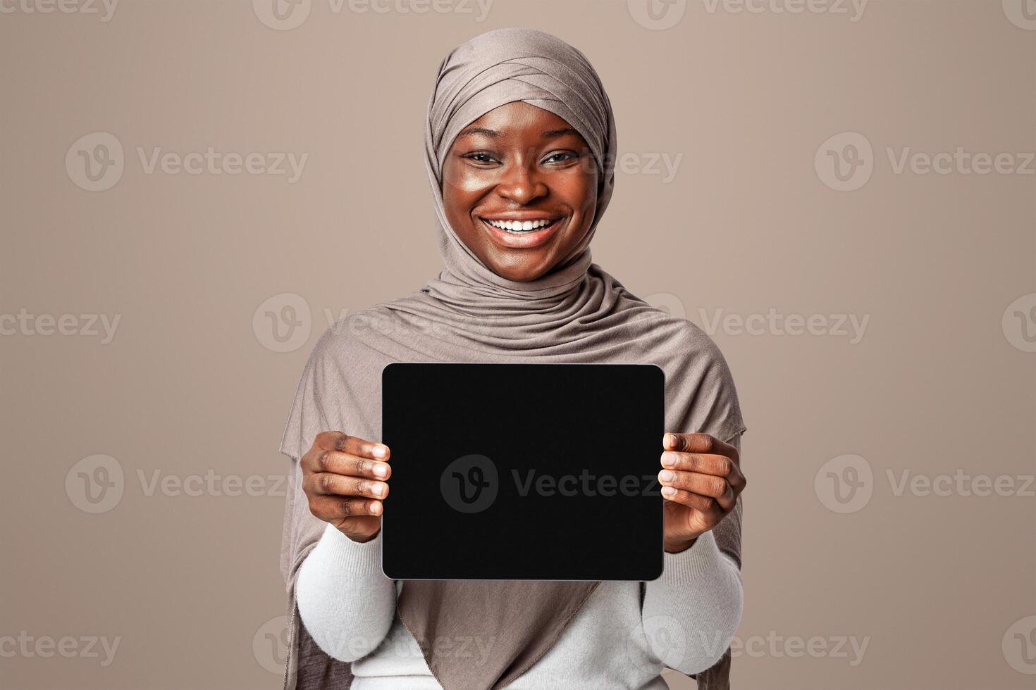 Mockup Image Of Smiling African Muslim Lady Holding Digital Tablet With Black Screen, Islamic Woman In Hijab Demonstrating Device With Copy Space For Your Website Or App Design Over Light Background photo
