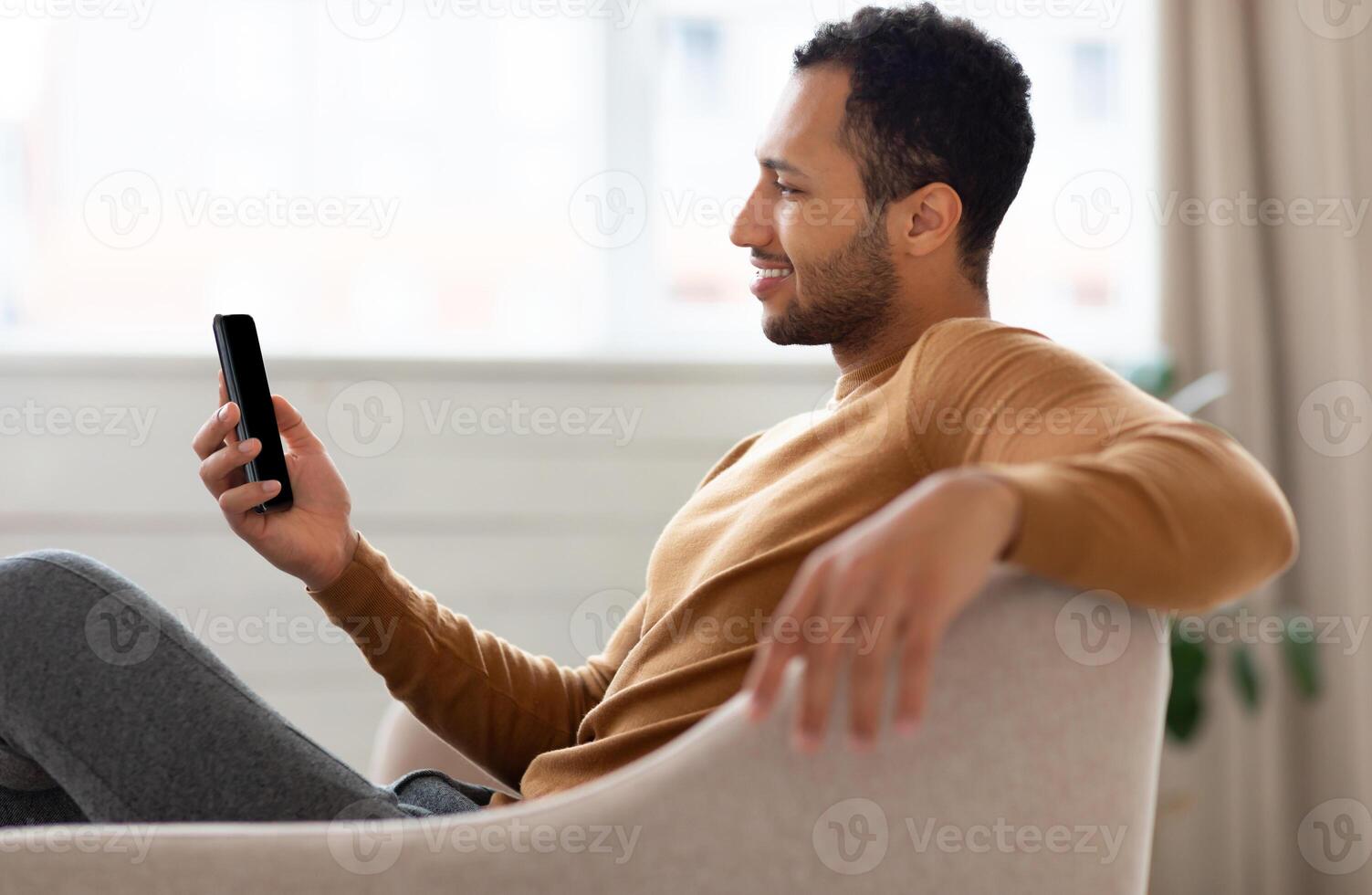 Profile side view portrait of young smiling Arab man holding mobile phone sitting on the couch in living room. Cool guy browsing internet, surfing web or watching , using application, free space photo
