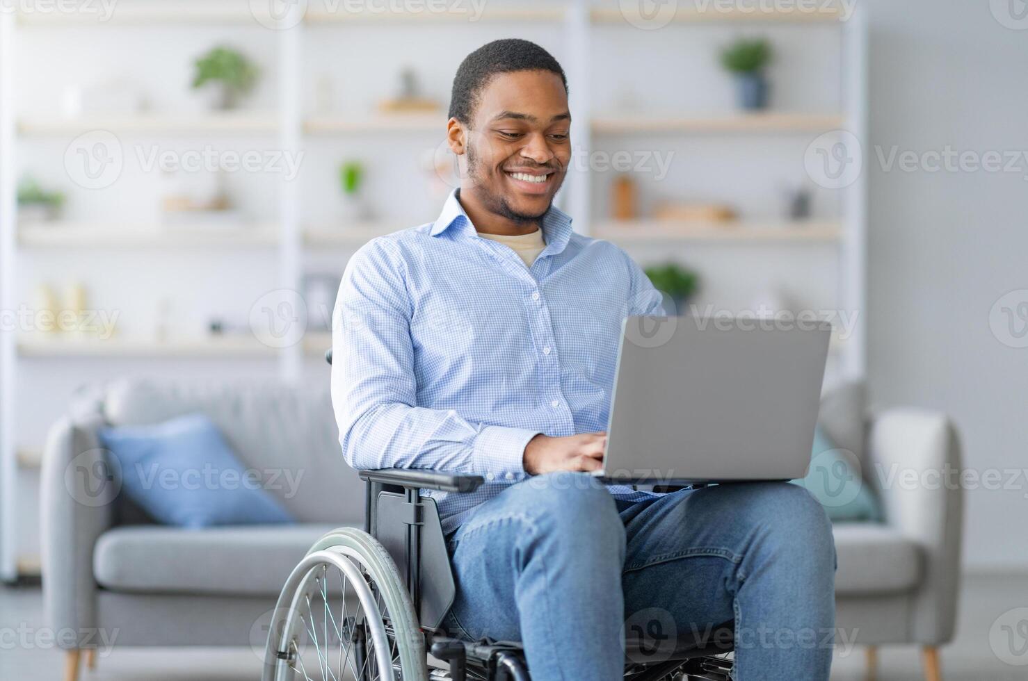 Portrait of cheerful disabled young black man in wheelchair using laptop for remote job at home. Happy black guy with physical disability having online work during covid pandemic photo