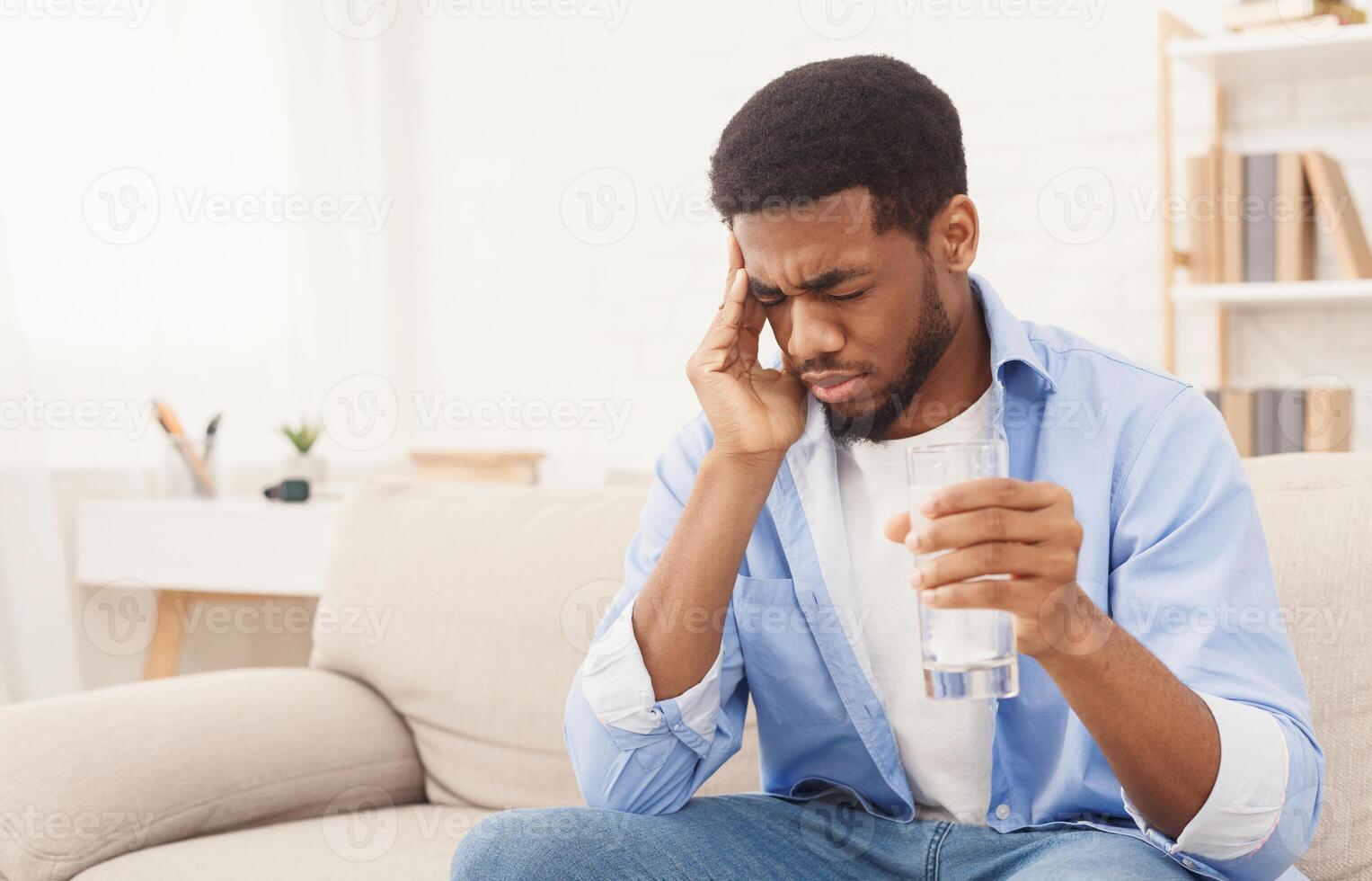 Exhausting headache. Sick african-american man taking pill with glass of water, suffering from migraine at home, copy space photo