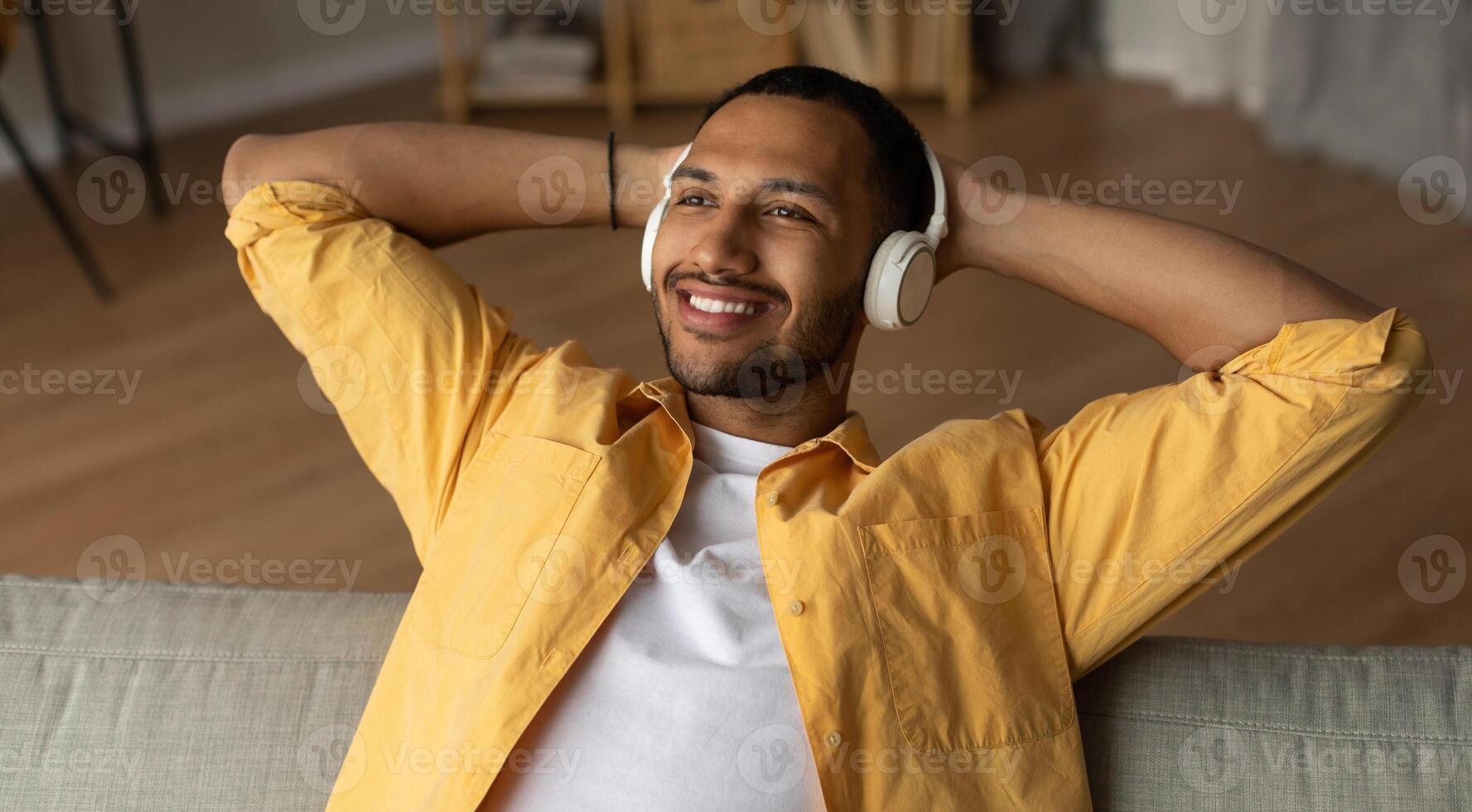 Handsome young black guy in headphones relaxing on couch, listening to music at home, banner design. Millennial African American man enjoying audio book in headset, resting indoors photo
