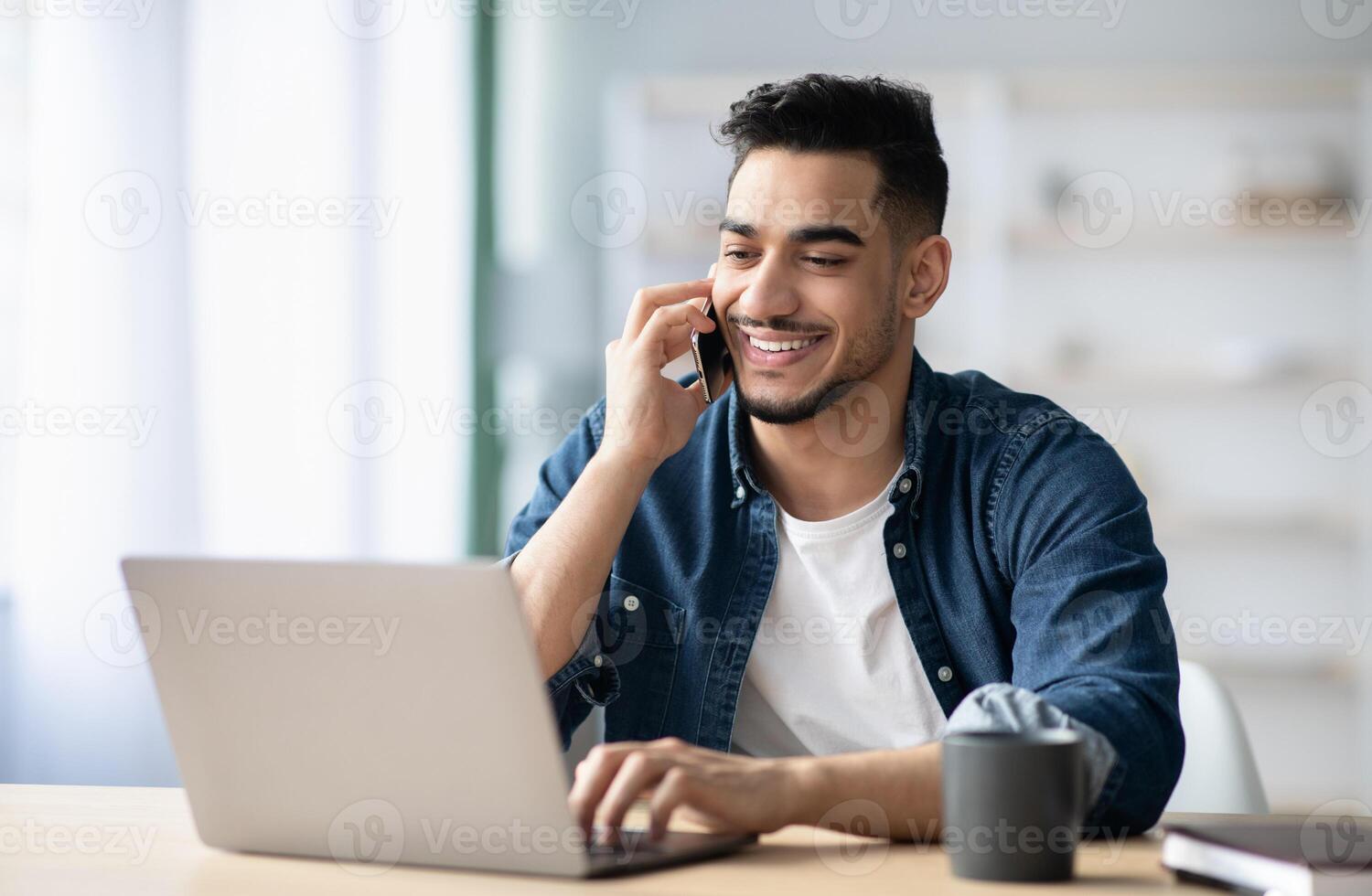 Successful middle-eastern guy talking on mobile phone with partners while working online at home, holding smartphone and looking at laptop, arab man freelancer having business call, copy space photo