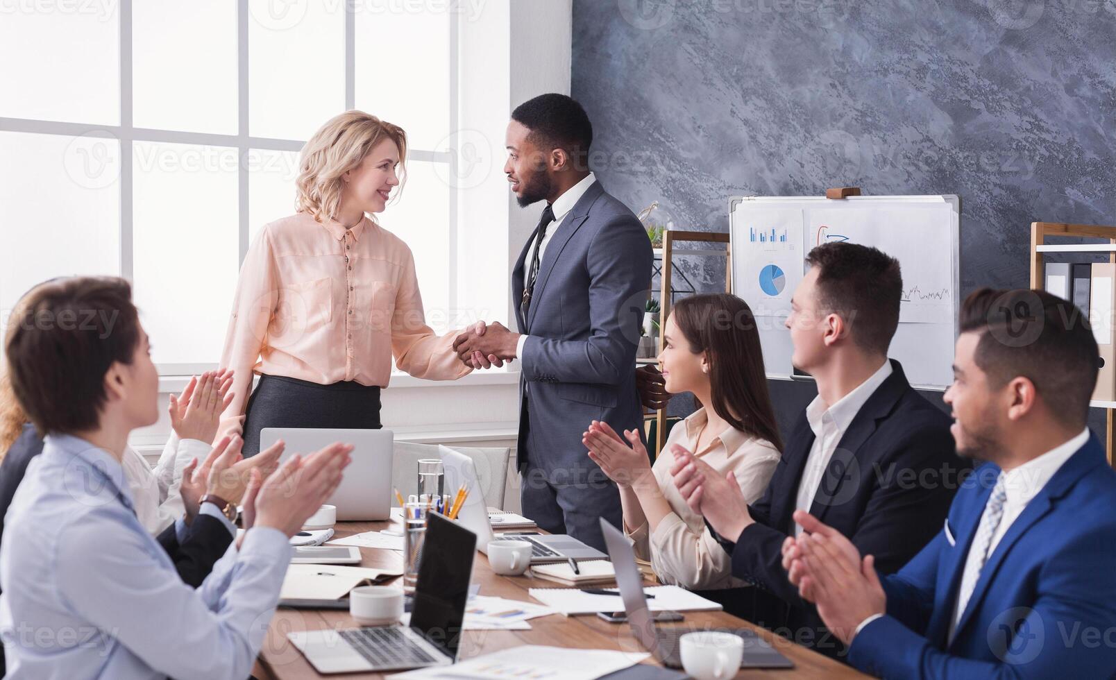 Group of cheerful business people sitting at meeting while boss and man shaking hands photo