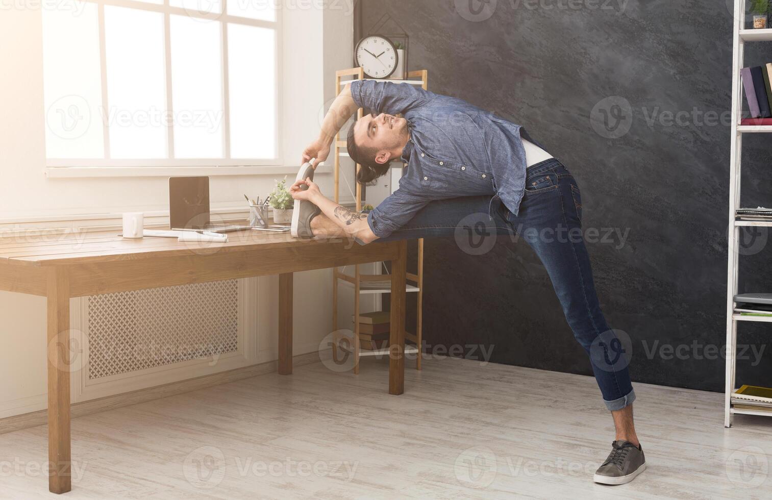Short break for yoga in office. Flexible man practicing yoga at workplace, making stretching exercise while using tablet, copy space. Active employee at work, healthy lifestyle concept photo