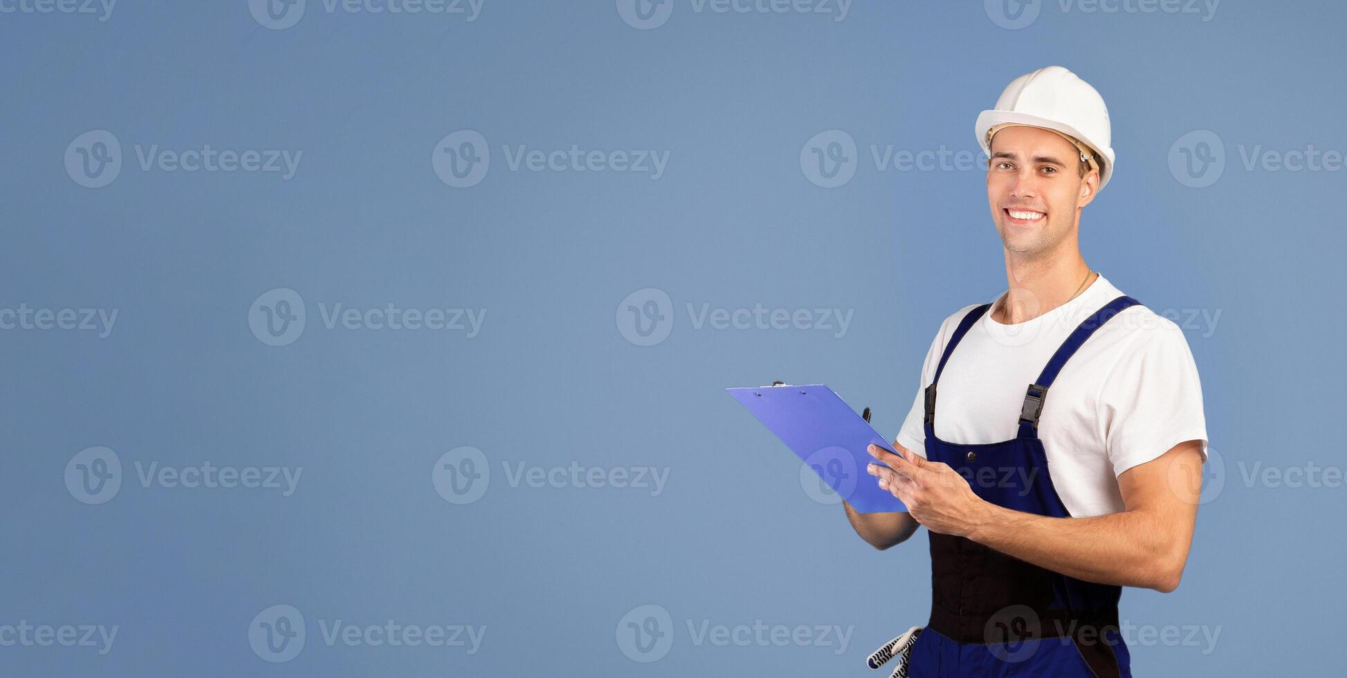 Repair And Renovation. Smiling handyman in hard hat helmet posing over blue background in studio, holding clipboard, taking notes, positive maintenance employee ready to work, looking at camera photo