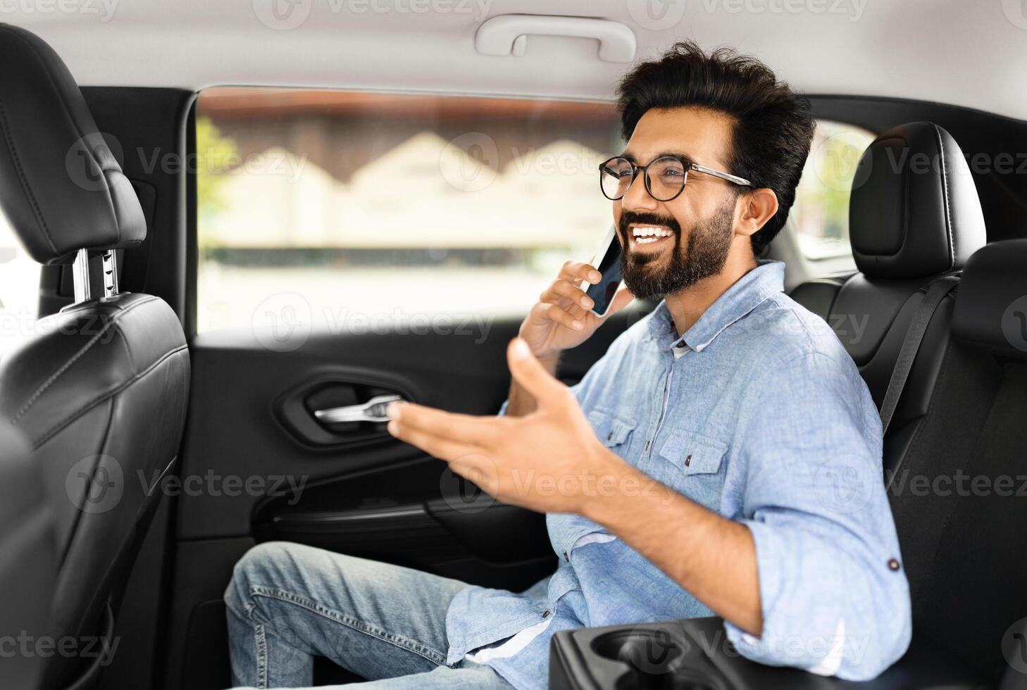 Positive young indian man talking over phone while sitting in taxi. Happy eastern guy sitting on back seat of car using mobile phone to communicate, talking over cellphone while commuting, going out photo