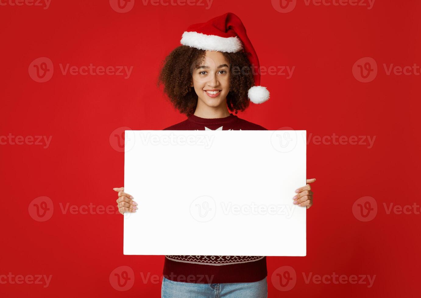 Merry Christmas and New Year mood. Cheerful beautiful young african american female in santa claus hat holds poster with blank space, isolated on red background, studio shot, cut out photo