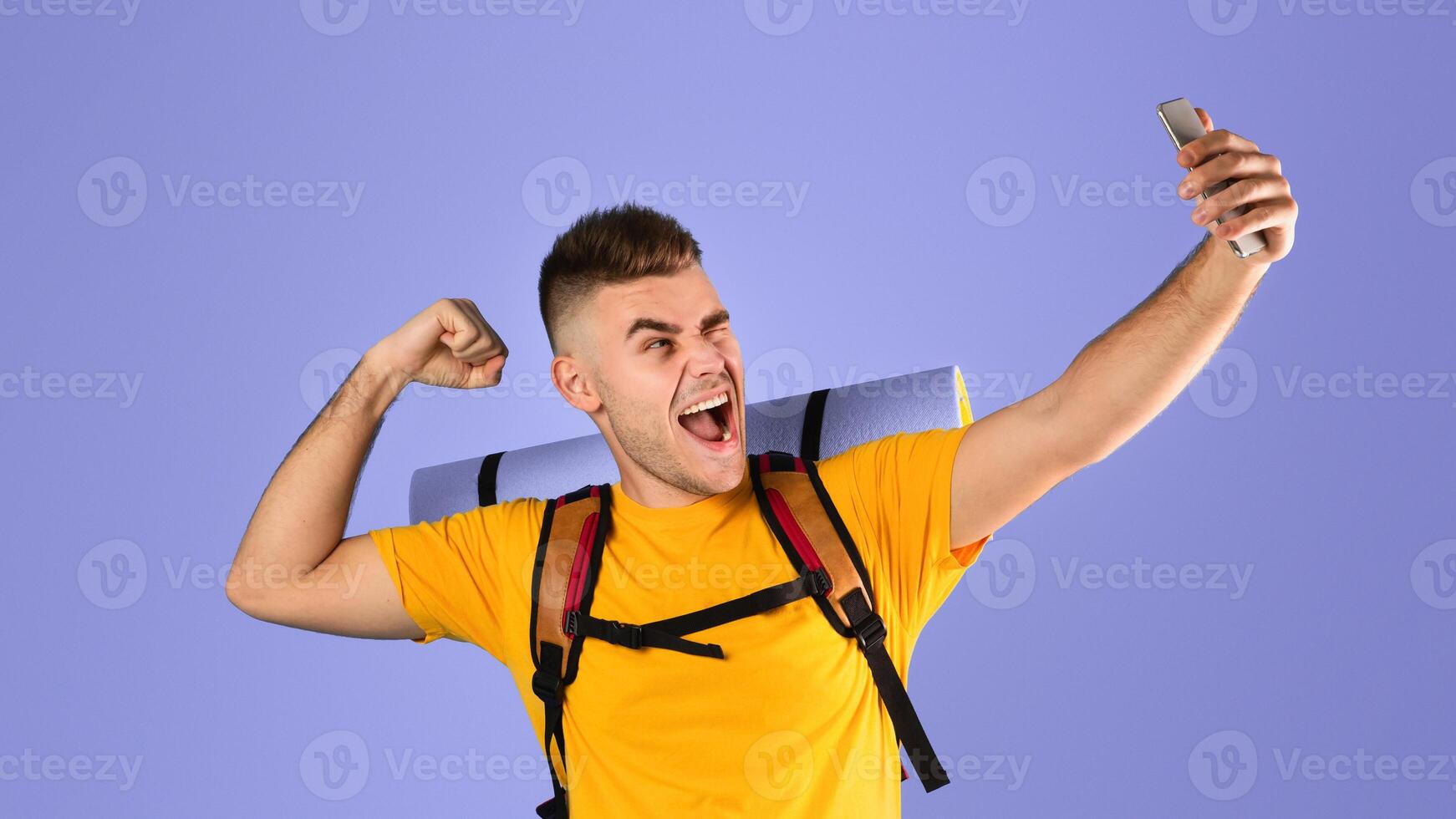 Cool young guy with tourist equipment taking selfie on smartphone over blue studio background. Funky millennial hiker making funny photo of himself on mobile device. Traveling adventure concept
