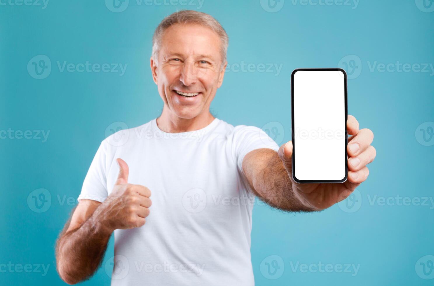 I Recommend. Portrait of smiling mature man holding smartphone with white blank screen in hand showing device and thumb up gesture. Selective focus on gadget with empty free space for mock up template photo