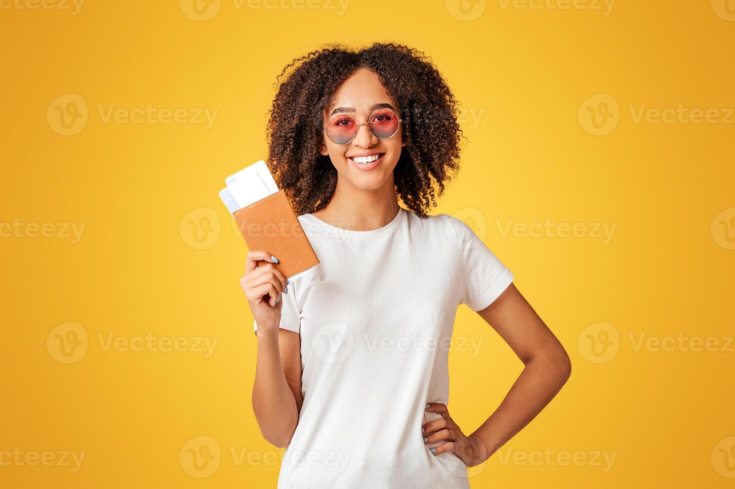 Low-cost, air travel and tourism. Happy young african american lady with curly hair in white t-shirt in glasses holds passport with air tickets isolated on yellow background, studio shot photo