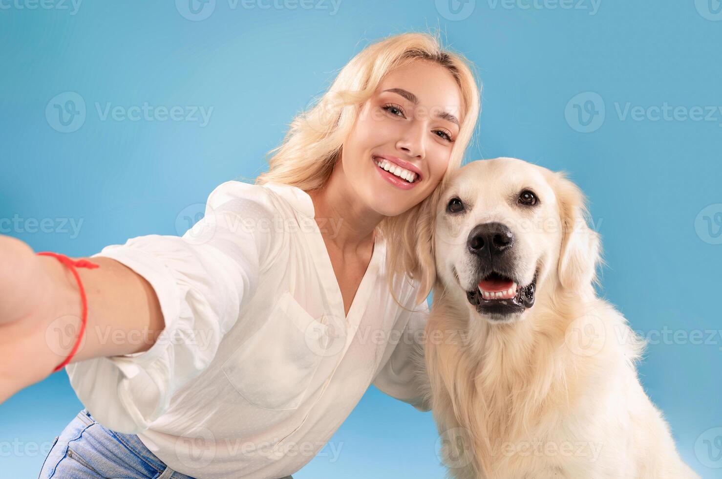 Friendship Concept. Portrait of excited beautiful caucasian blonde woman taking selfie with her happy dog, posing looking at camera, isolated on blue studio background wall, closeup photo
