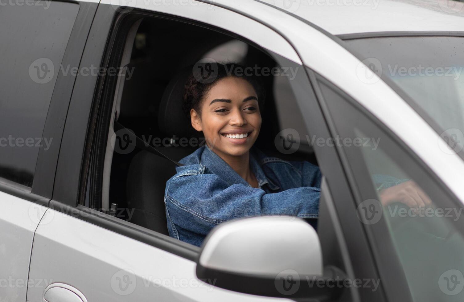 Woman with positive facial expression drives new car and enjoy auto trip. Young cheerful millennial african american female holds steering wheel, sits inside and looking out open window, empty space photo