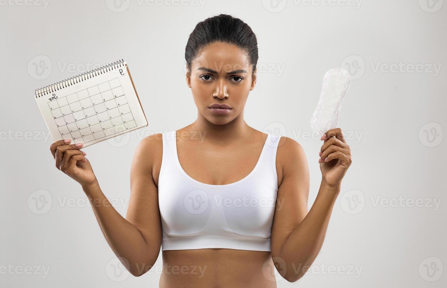Angry unhappy young black woman demonstrates periods calendar and sanitary napkin, isolated on grey studio background. Women healthcare, menstruation, menstrual cycle, period delay concept photo