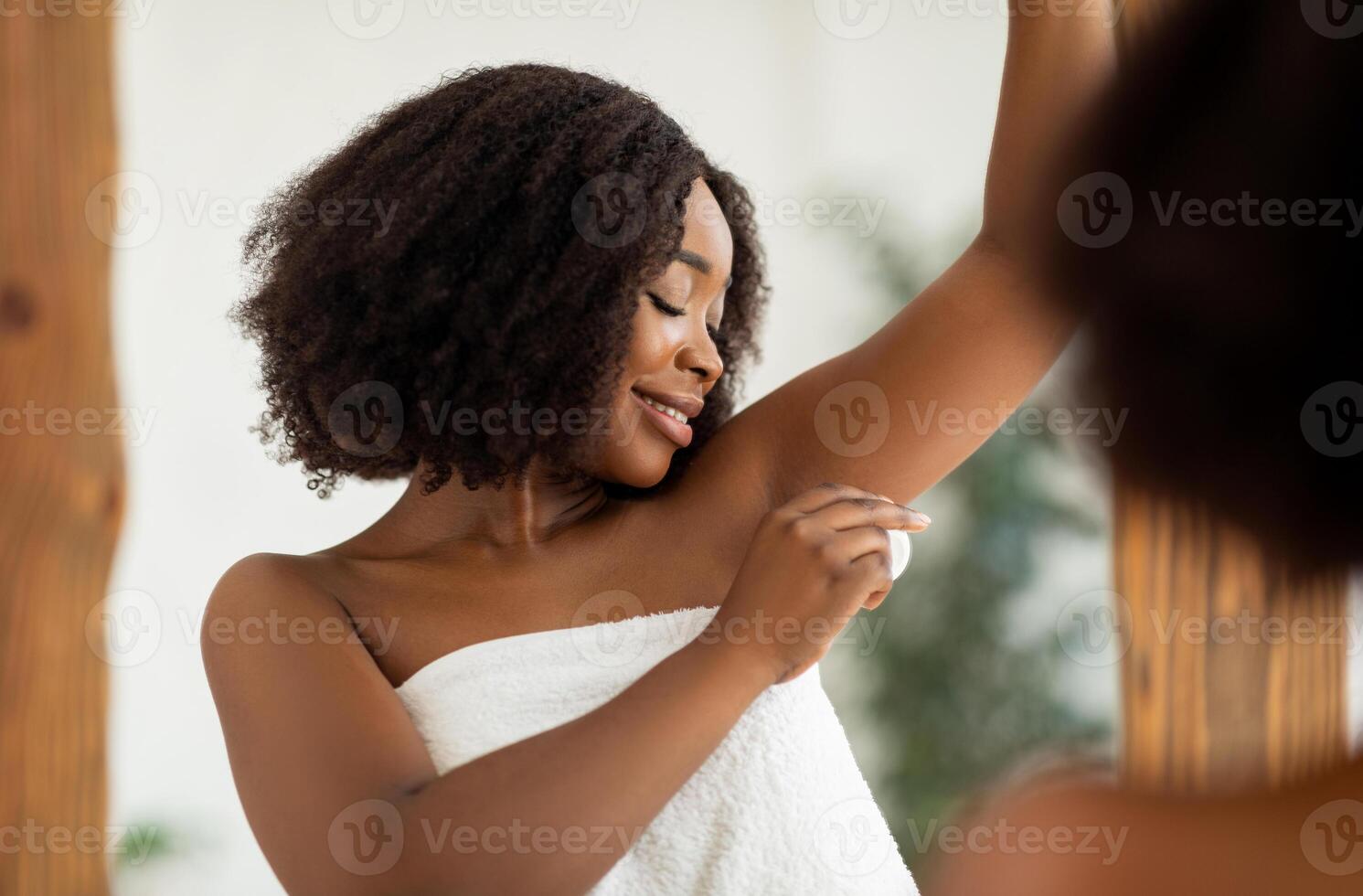 Portrait of pretty black woman applying deodorant after shower, standing in front of mirror at home. Millennial African American lady using antiperspirant to control sweating in summer photo