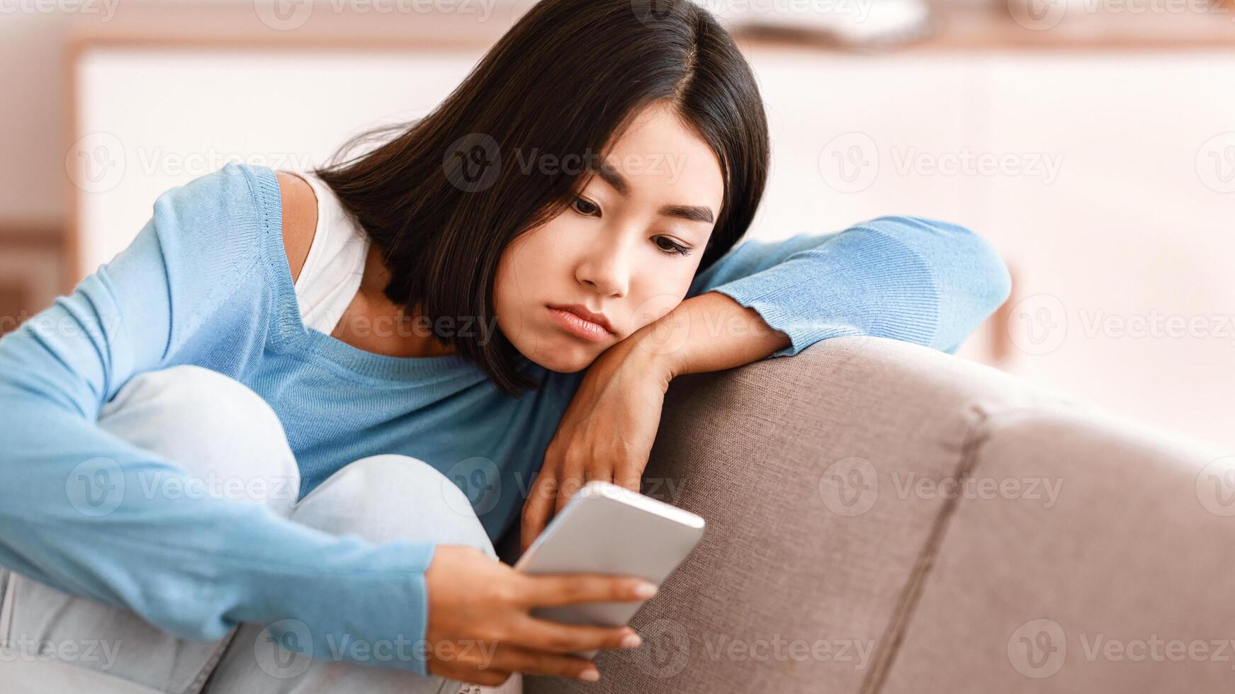 Depression Concept. Portrait of frustrated asian young woman looking at smartphone, checking social media, lying on the couch. Upset lady waiting for his message or call, reading bad news photo