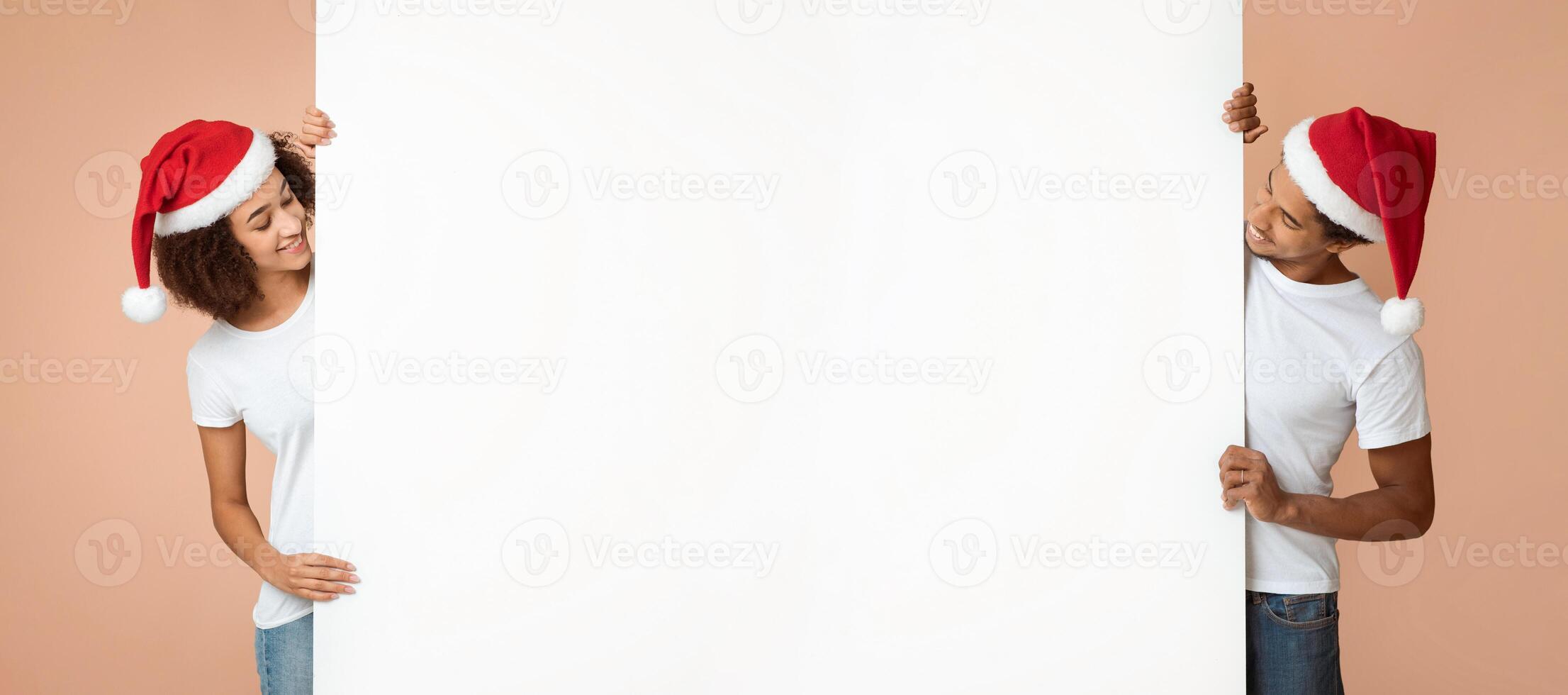 Happy together and Merry Xmas together. Smiling millennial african american man and woman in Santa Claus hats look at each other from behind white blank banner for advert isolated on light background photo