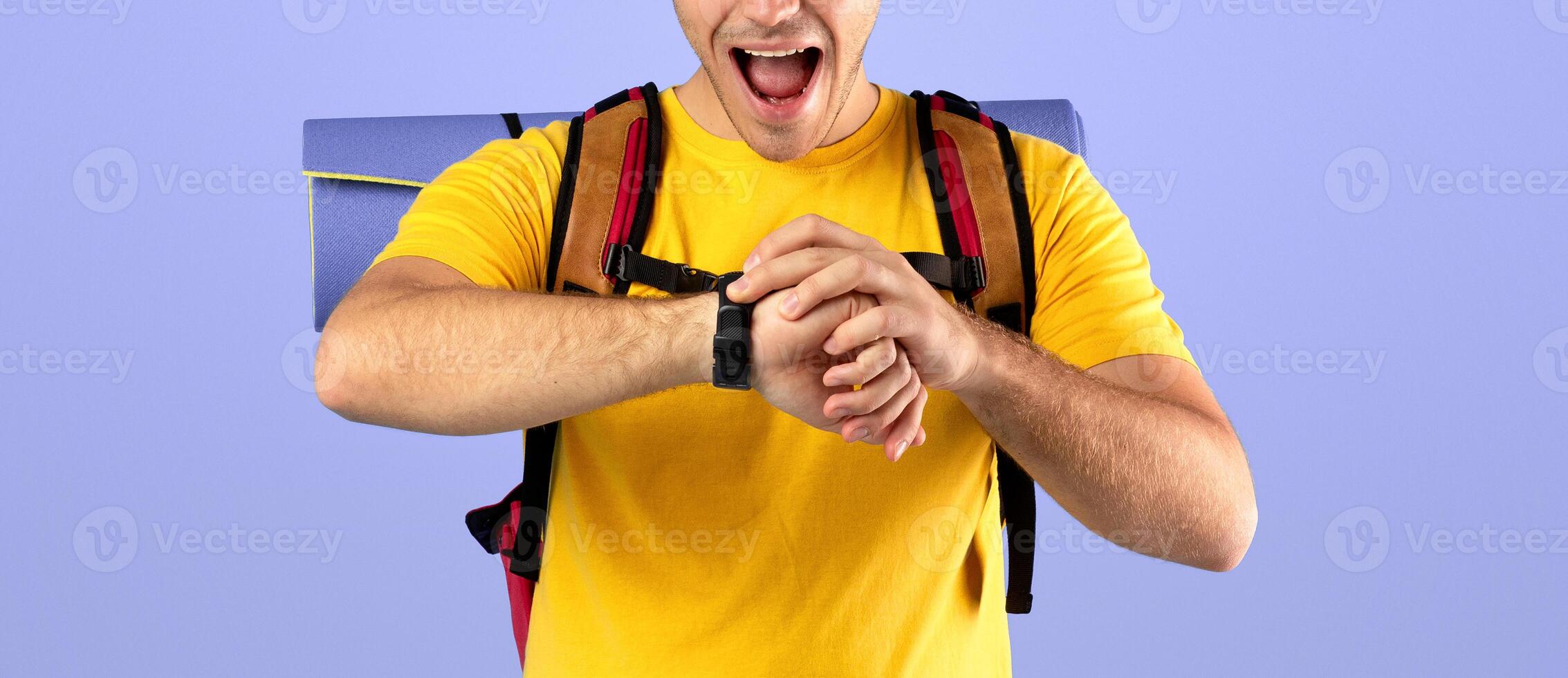 Worried young guy with camping equipment looking at his watch, checking time over blue studio background. Shocked millennial hipster missing his ride, late for bus or airplane photo