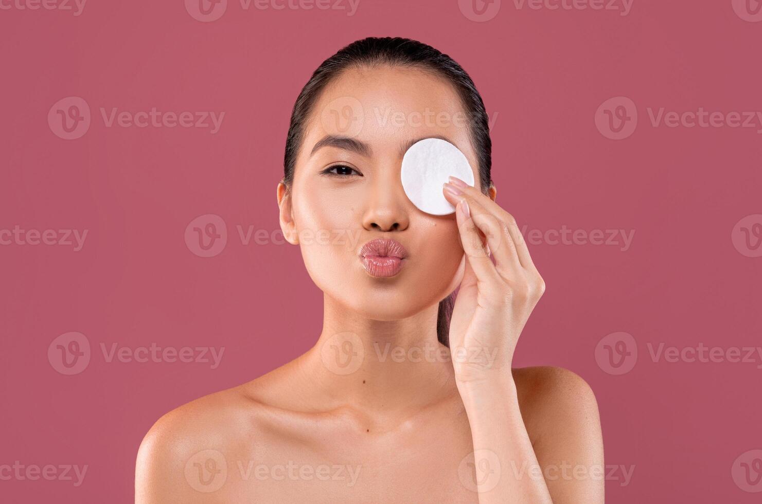 Closeup photo of happy topless young korean woman cleansing her eyes with cotton pad and smiling over purple studio background, removing makeup with beauty product, showing kissing lips, copy space