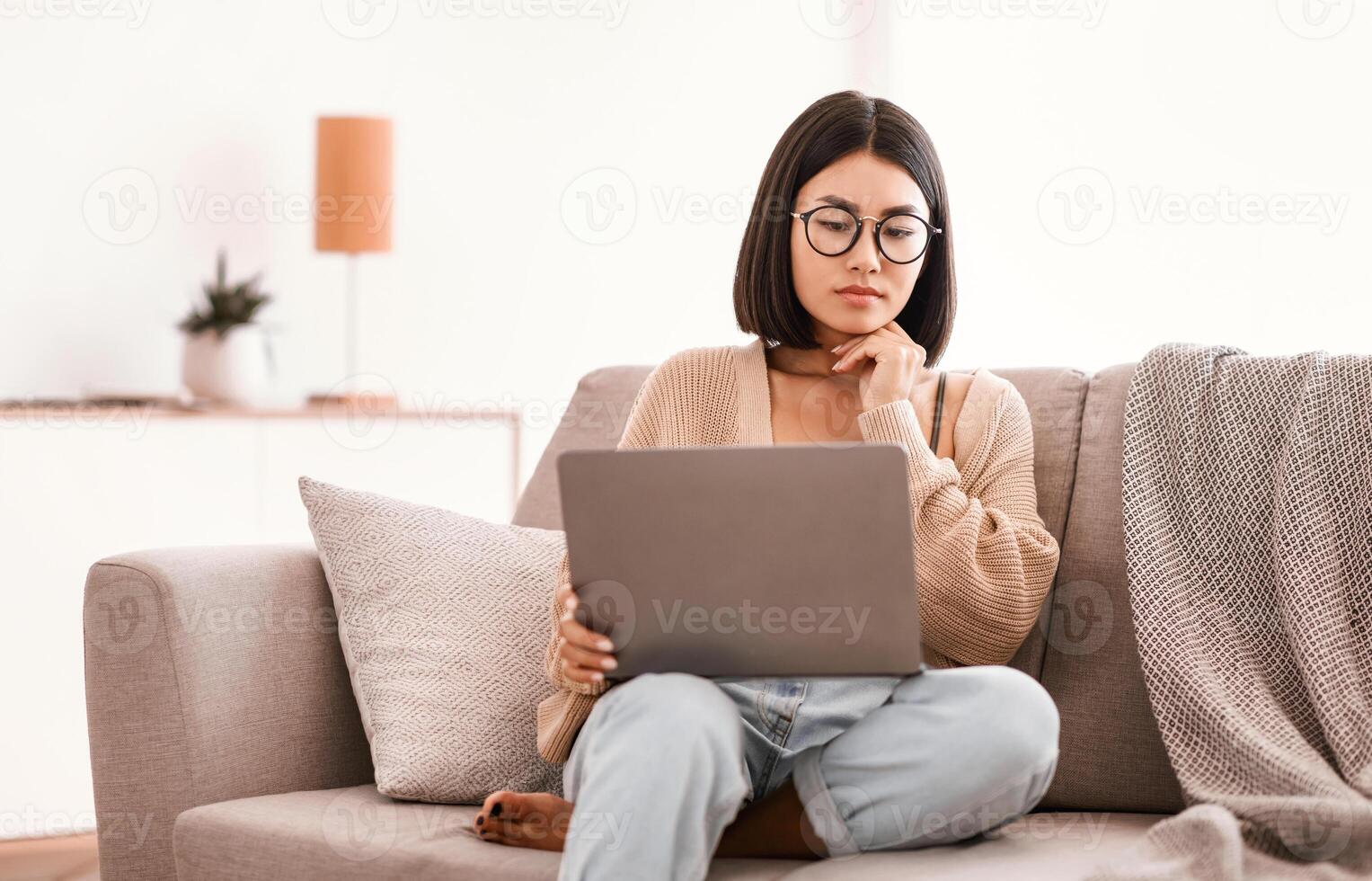 Pensive asian female freelancer in glasses with laptop brainstorming at new project, making research in internet, having remote job, sitting on couch in living room at home, looking at computer screen photo