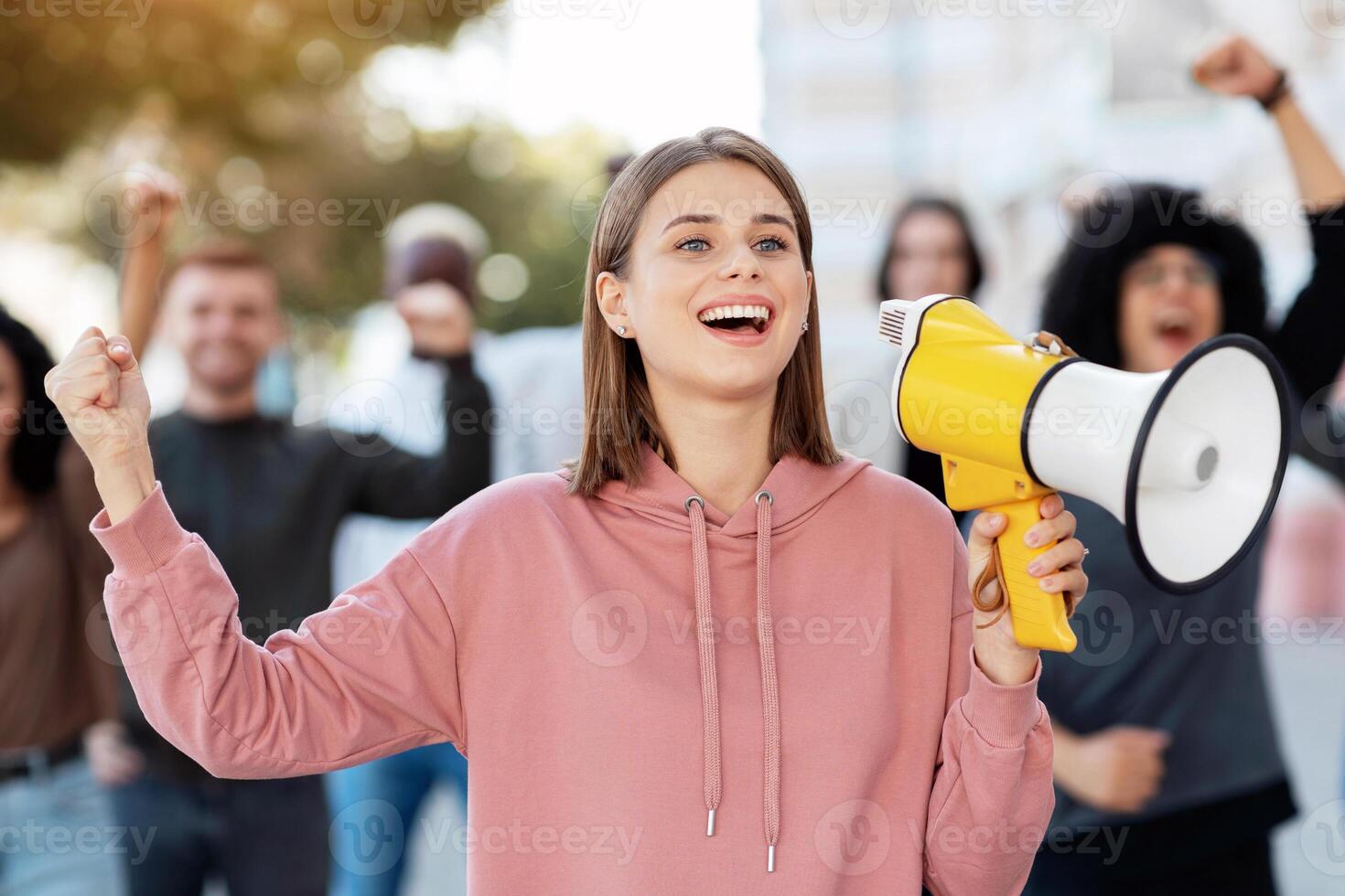 Enthusiastic blonde lady activist with megaphone on the street, chanting slogans and raising fist up, cheerful young woman leading international group of activists, striking against lockdown rules photo