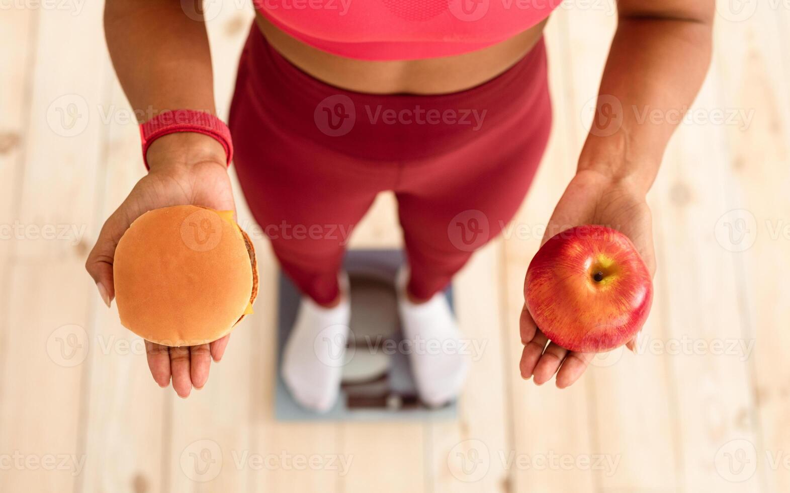 Healthy Diet And Weight Loss. Slim Black Girl Choosing Between Burger And Apple Standing On Weight-Scales At Home. High-Angle, Cropped photo