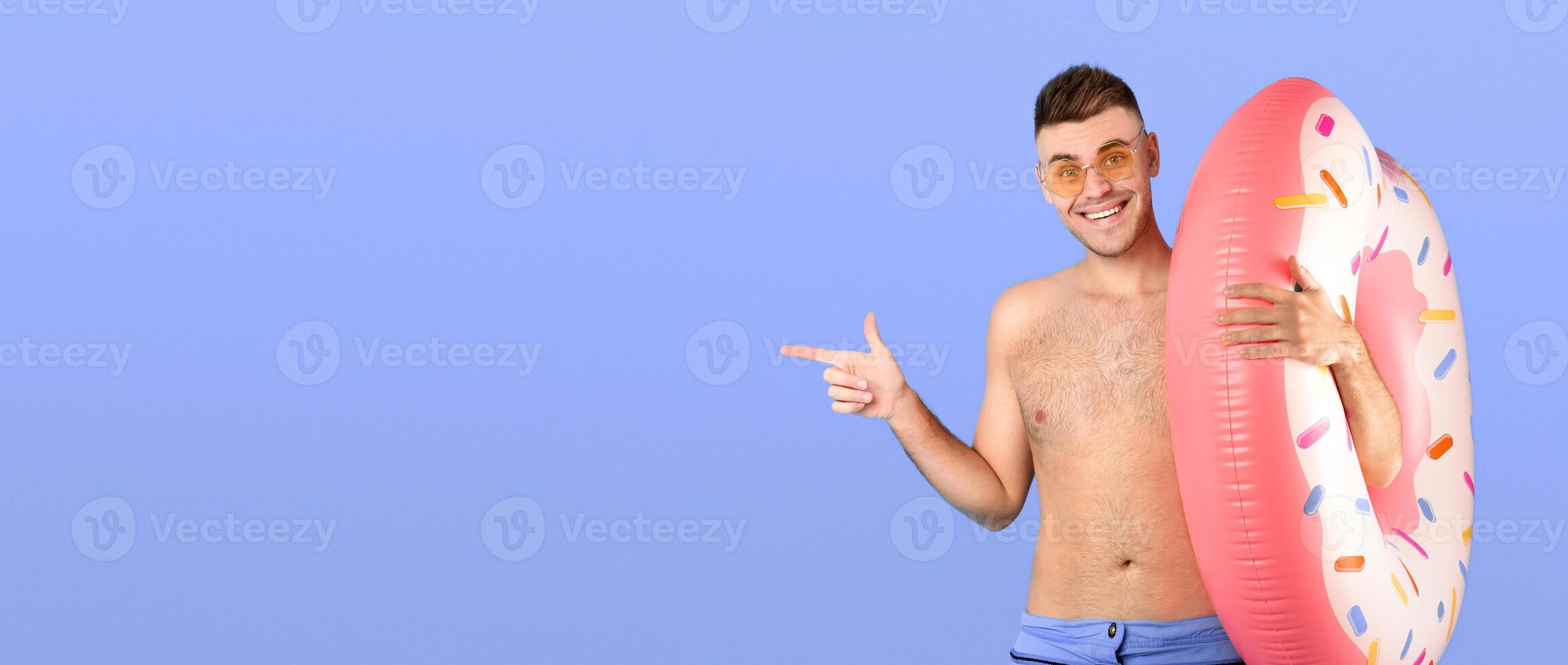 Smiling young guy in shorts with inflatable ring pointing aside at empty space over blue studio background, banner design. Attractive millennial man planning summer vacation in tropical country photo