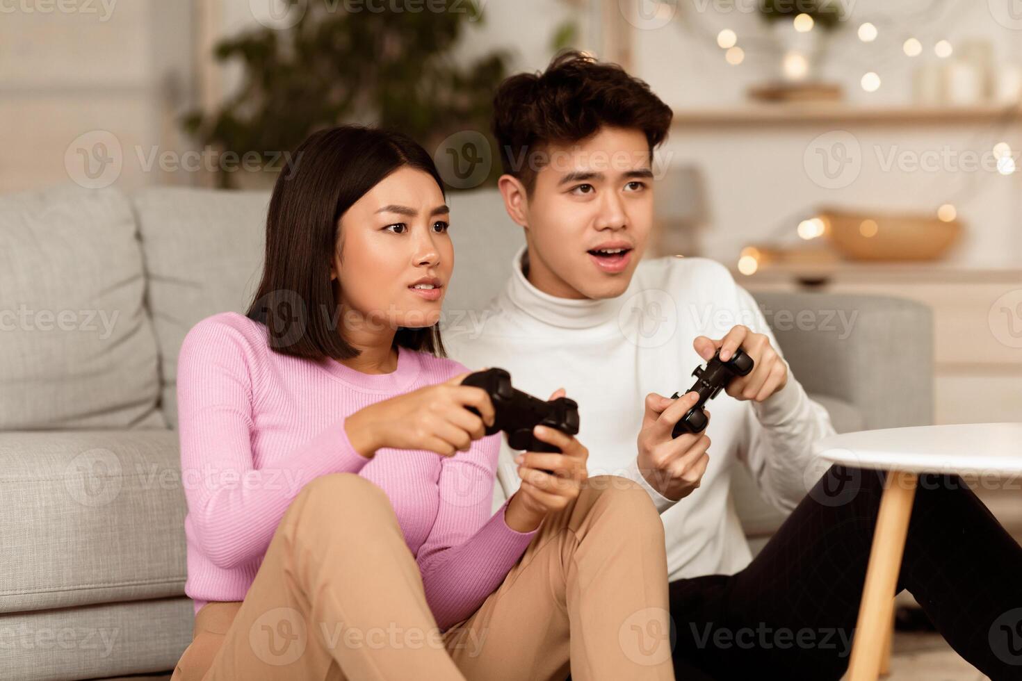 Chinese Millennial Couple Playing Computer Game Holding Joysticks Sitting On Floor At Home Having Fun On Weekend. Asian Boyfriend And Girlfriend Play Videogame In The Evening photo