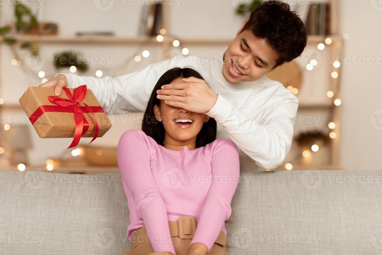 New Year Celebration. Asian Husband Giving Christmas Gift To Wife, Covering Her Eyes Celebrating Holiday Sitting On Coush At Home. Birthday And Xmas Presents, Happy Winter Holidays Concept photo