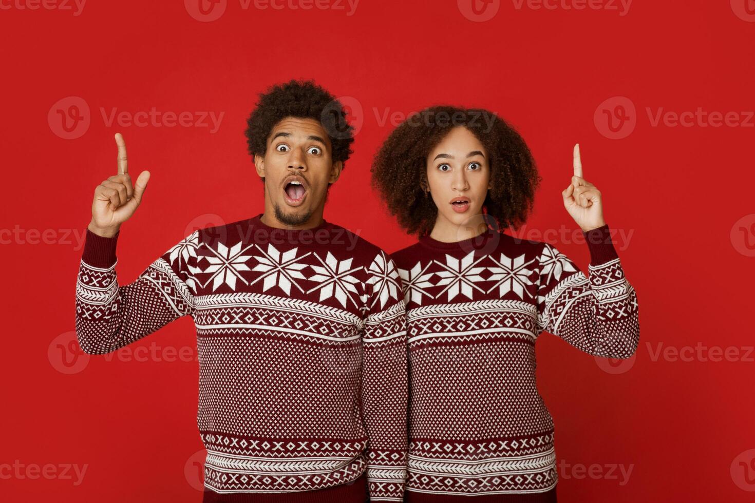 Surprise and shock, natural human emotions. Amazed young african american man and lady in christmas sweaters with open mouths, points fingers up to empty space, isolated on red background, studio shot photo