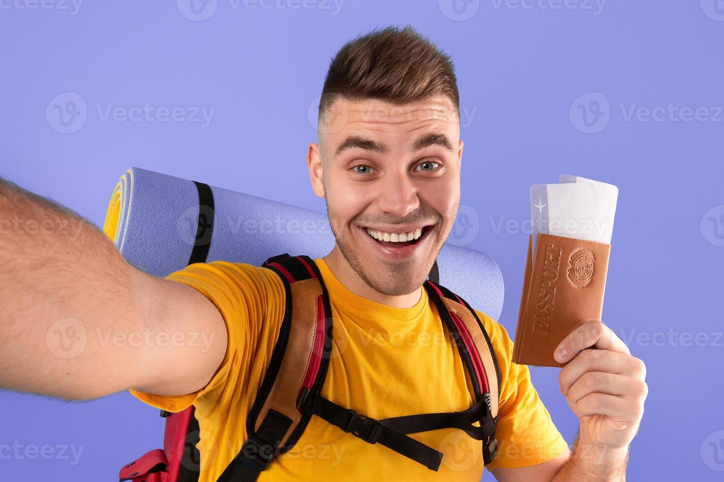 Happy young guy with backpack, passport and plane tickets taking selfie on blue background photo
