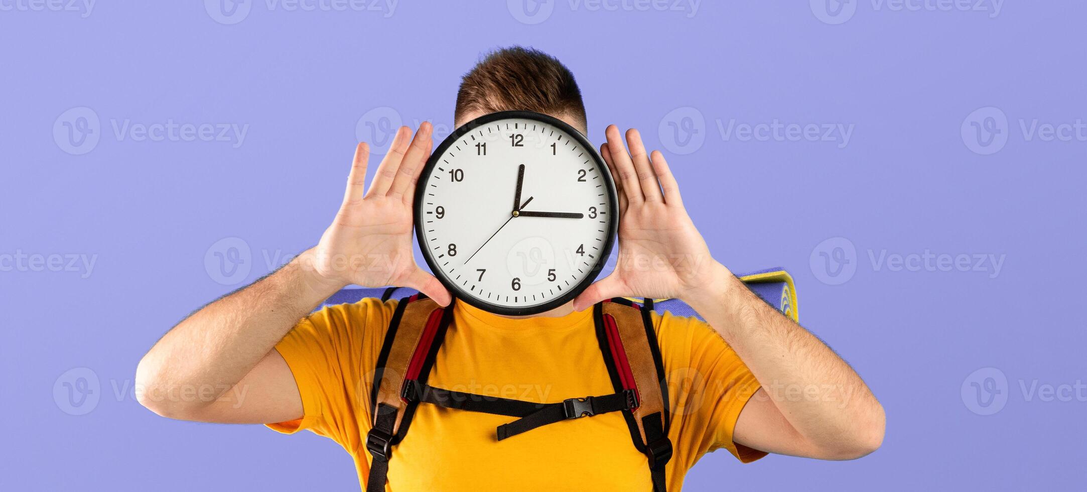 Time to travel concept. Young man with camping equipment holding clock in front of his face over blue studio background. Millennial guy ready to start his hiking journey or vacation photo