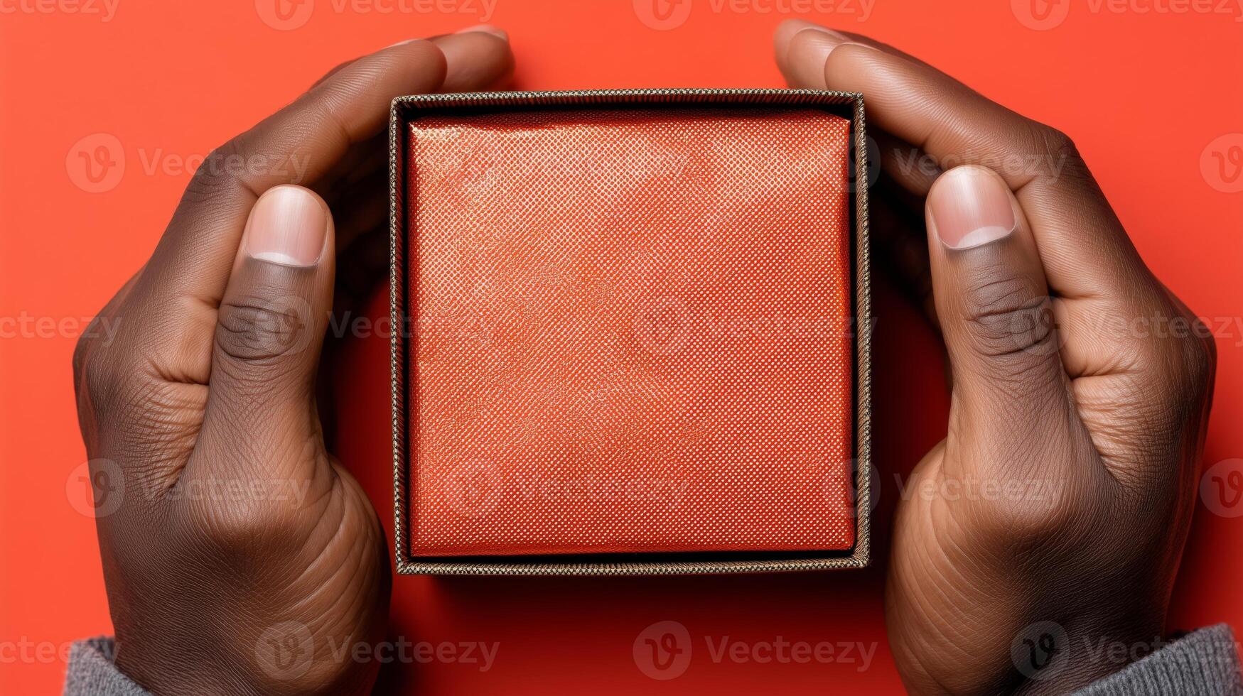 Two hands gently grasp an open box, showcasing its textured exterior. The box rests on a vivid red surface, highlighting its warm color. This moment captures the anticipation of discovery. photo
